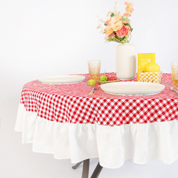 Round table with red and white checkered tablecloth, plates, glasses, and a vase with flowers on a white background.