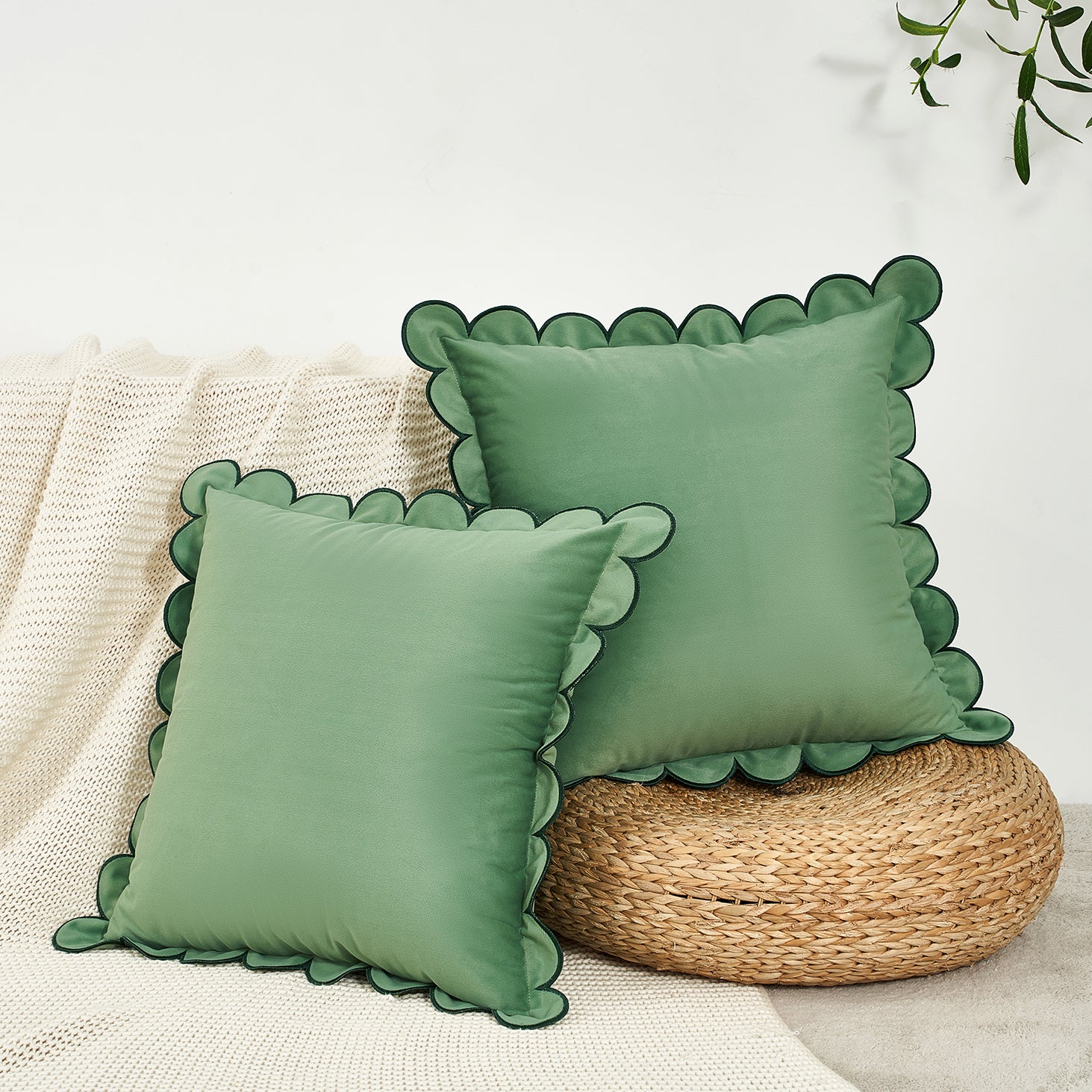 Two green pillows with scalloped edges on a woven stool against a neutral background.