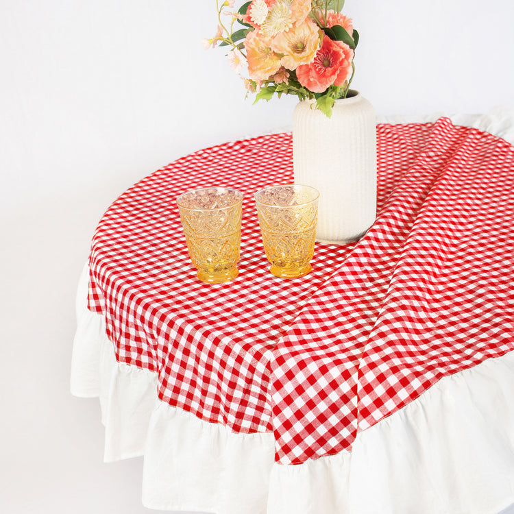 Round table with red and white checkered tablecloth, two yellow glasses, and a vase with flowers on a white background.