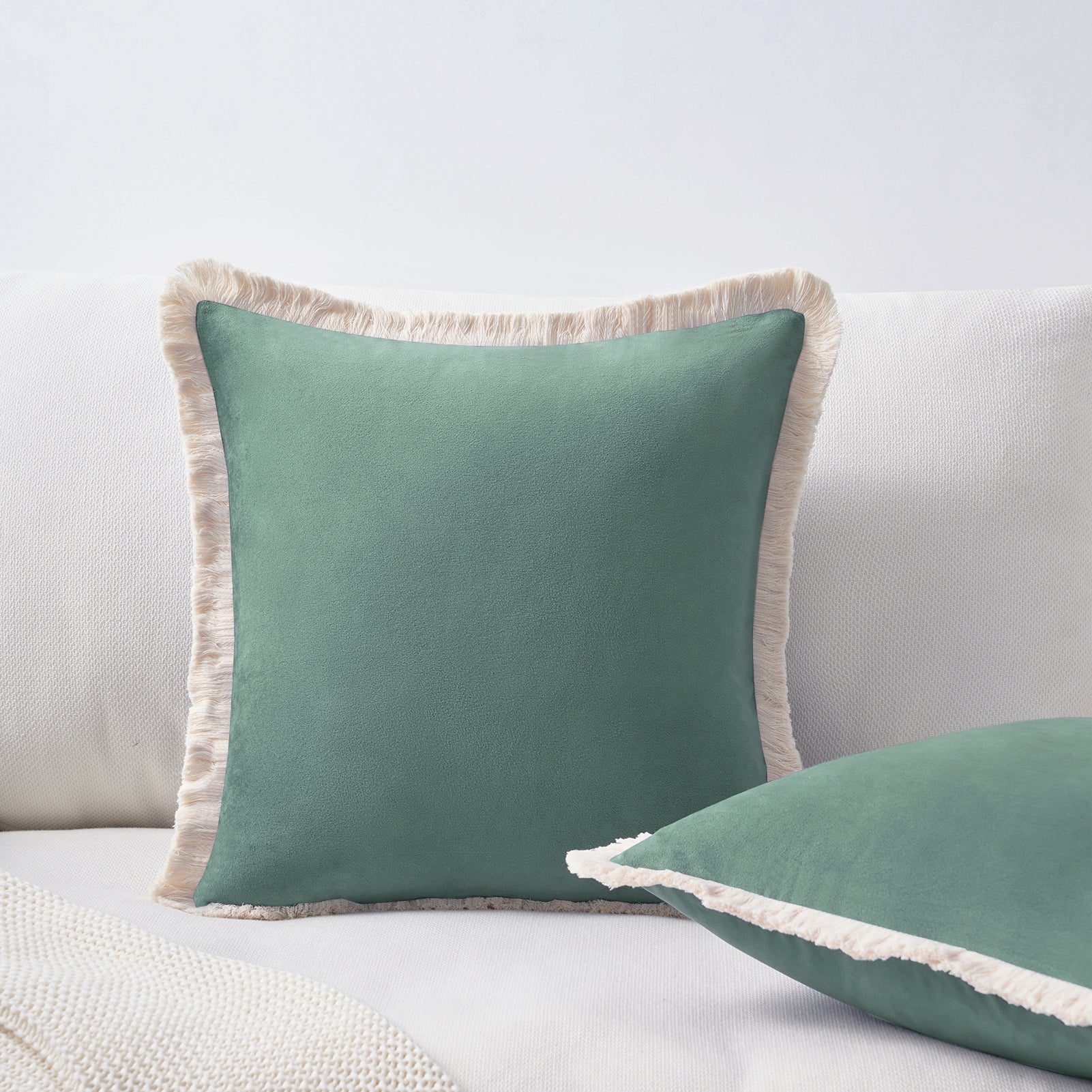 Two green pillows with beige fringes on a light gray sofa.