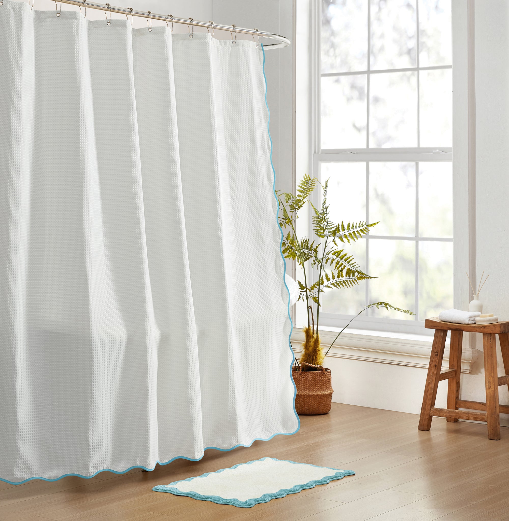White shower curtain with blue trim in a bathroom setting with a plant and stool.