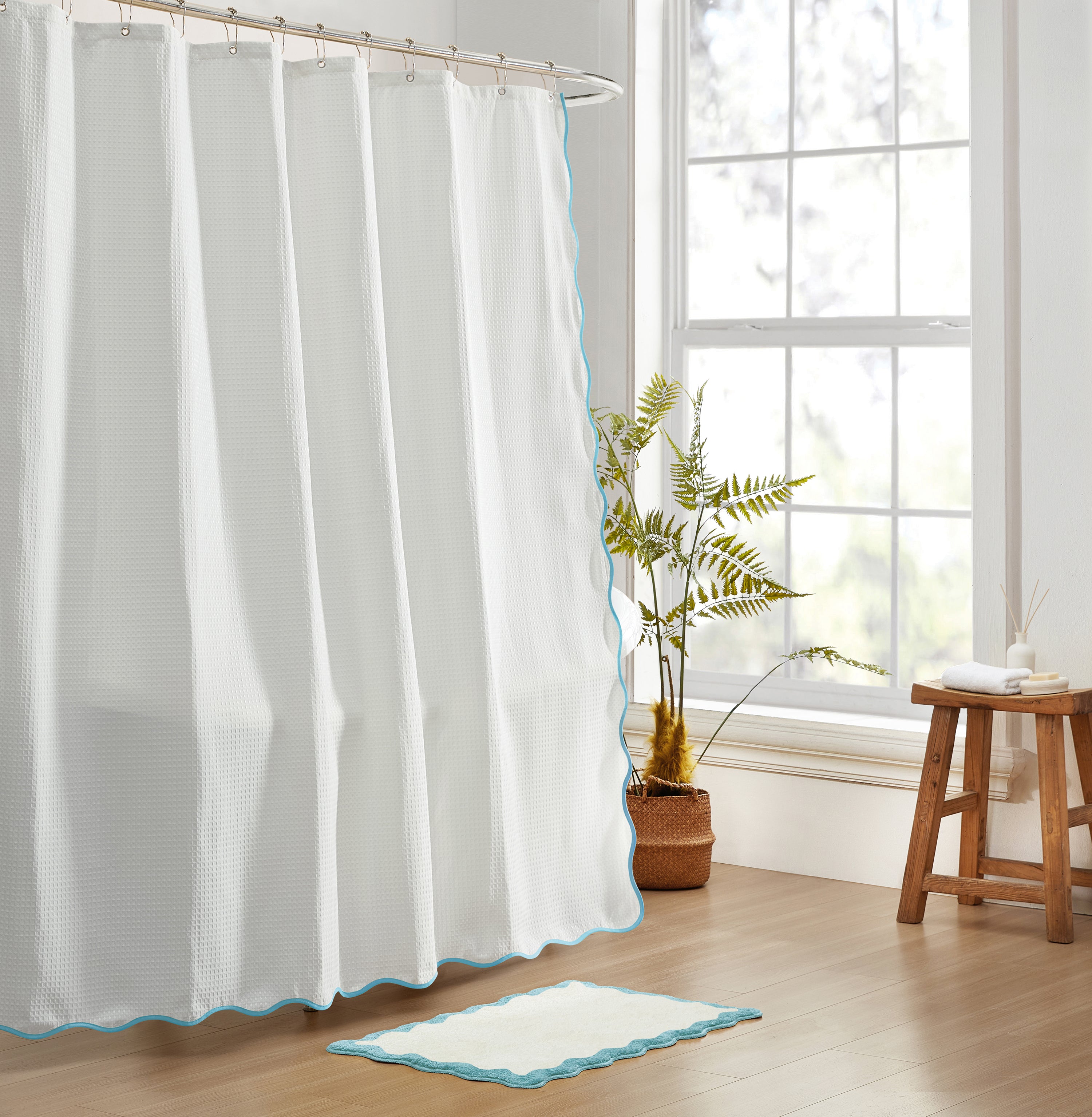 White shower curtain with blue trim in a bathroom setting with a plant and stool.