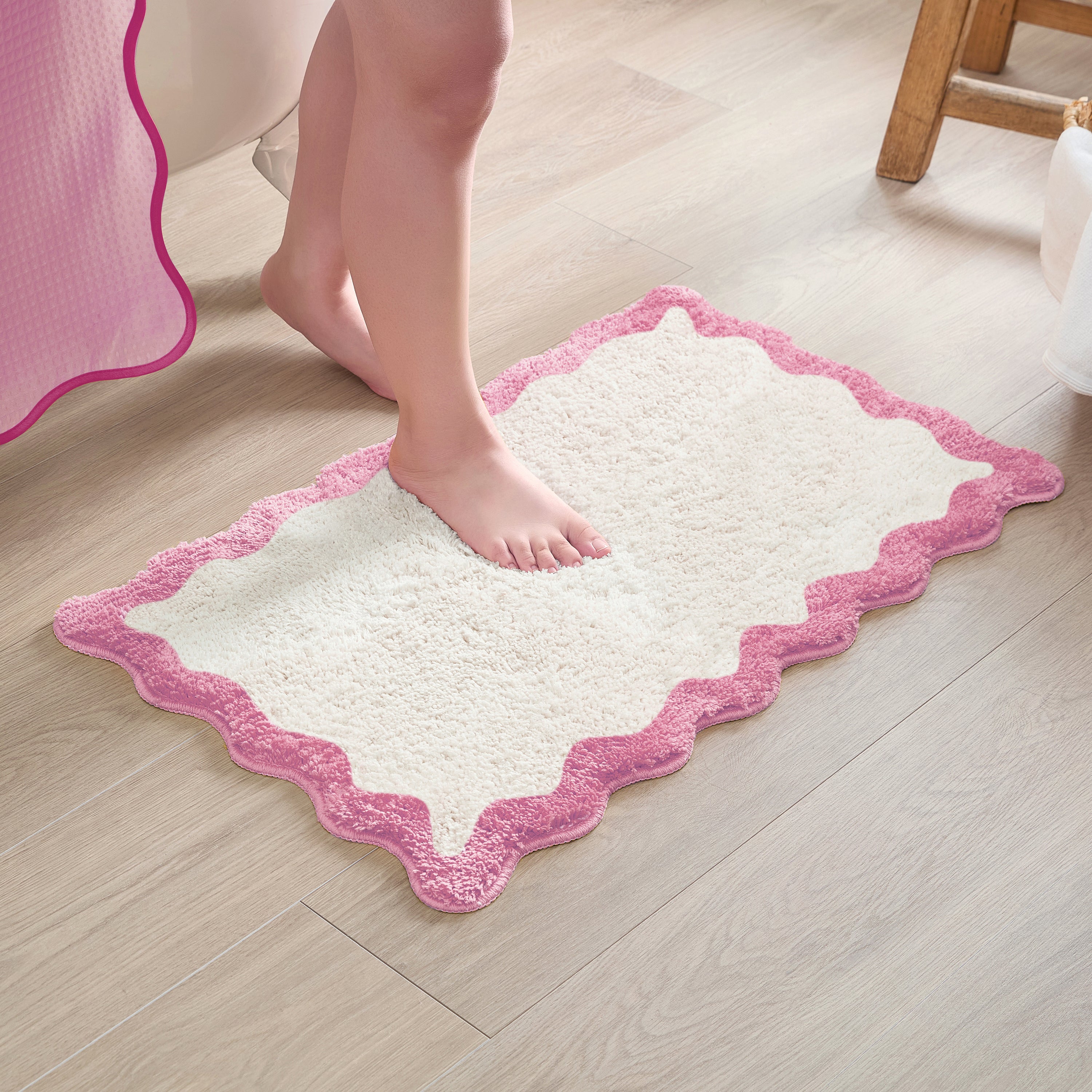 White bath mat with pink border on a wooden floor, person stepping onto it.