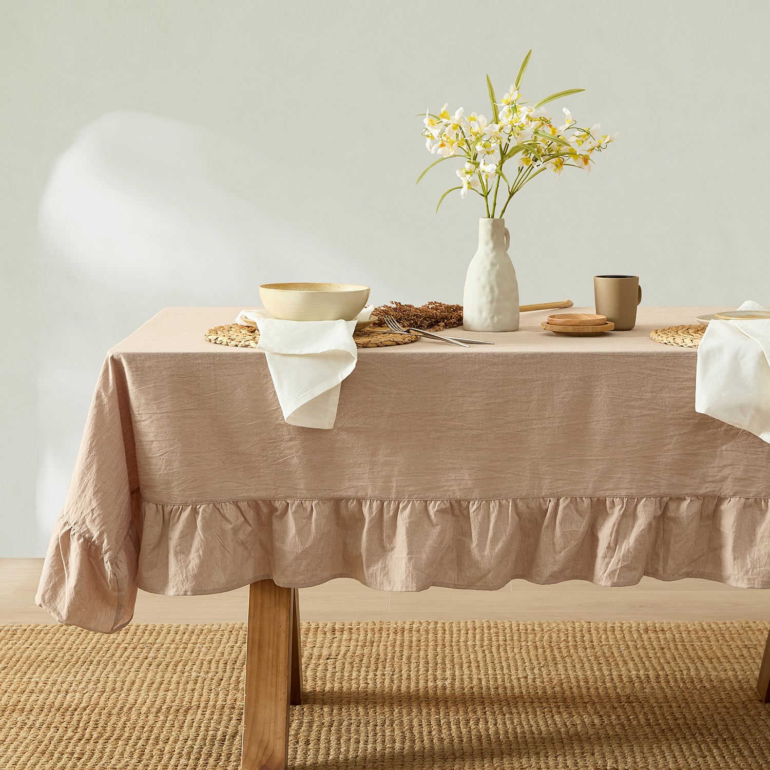 Beige tablecloth on a wooden table with a vase of flowers and other decor items.