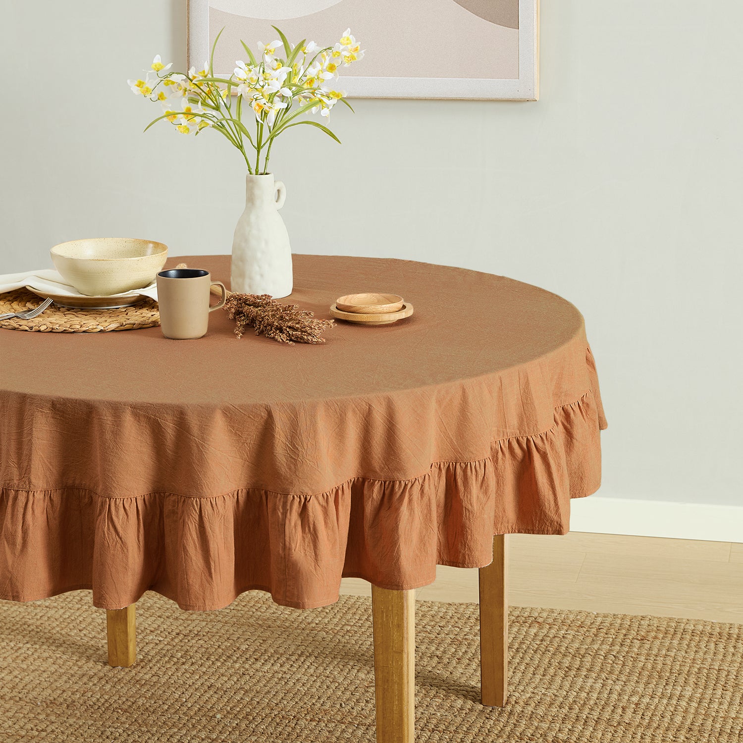Round table with a brown tablecloth, mug, plates, and vase with flowers in a room.