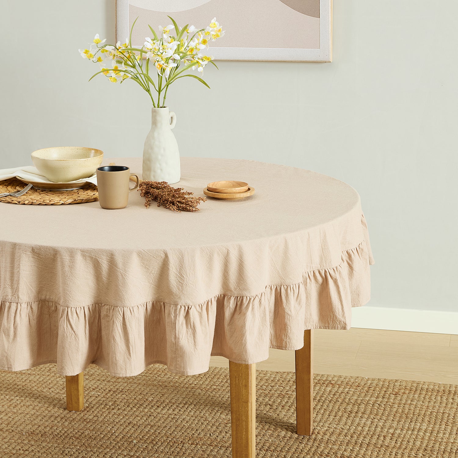 Round table with a beige tablecloth, mug, vase with flowers, and plates on a light green wall background.
