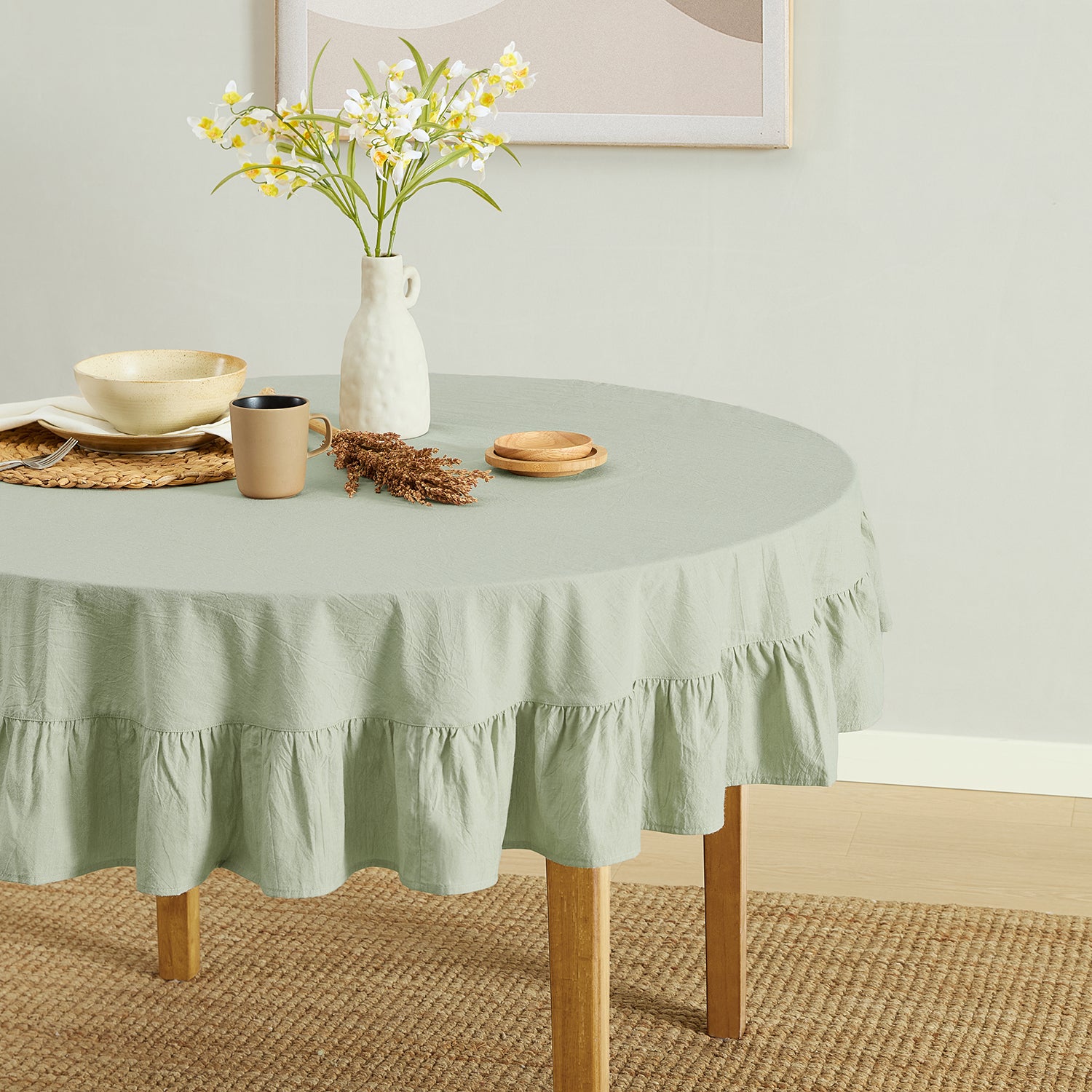 Round table with a green tablecloth, mug, vase with flowers, and plates on a light green wall background.
