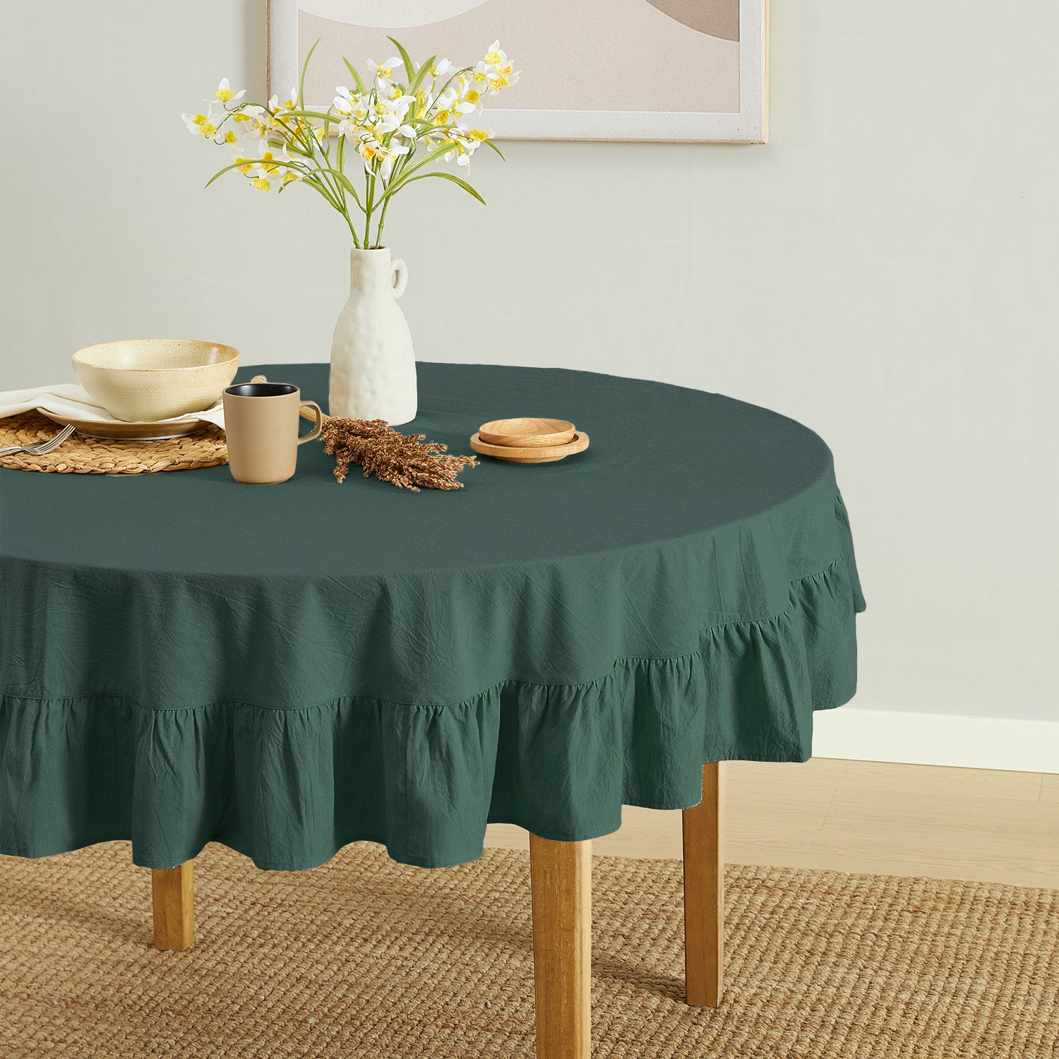 Round table with a green tablecloth, mug, vase, and flowers in a room.