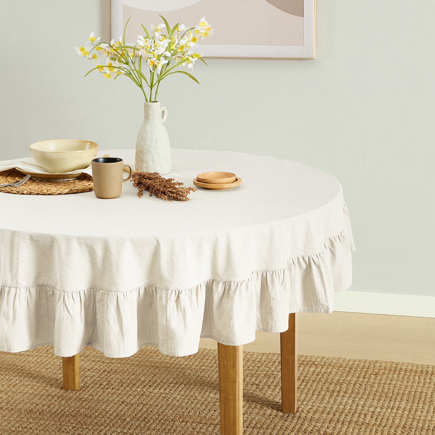 Round table with a white tablecloth, mug, vase with flowers, and decorative items on a neutral background.