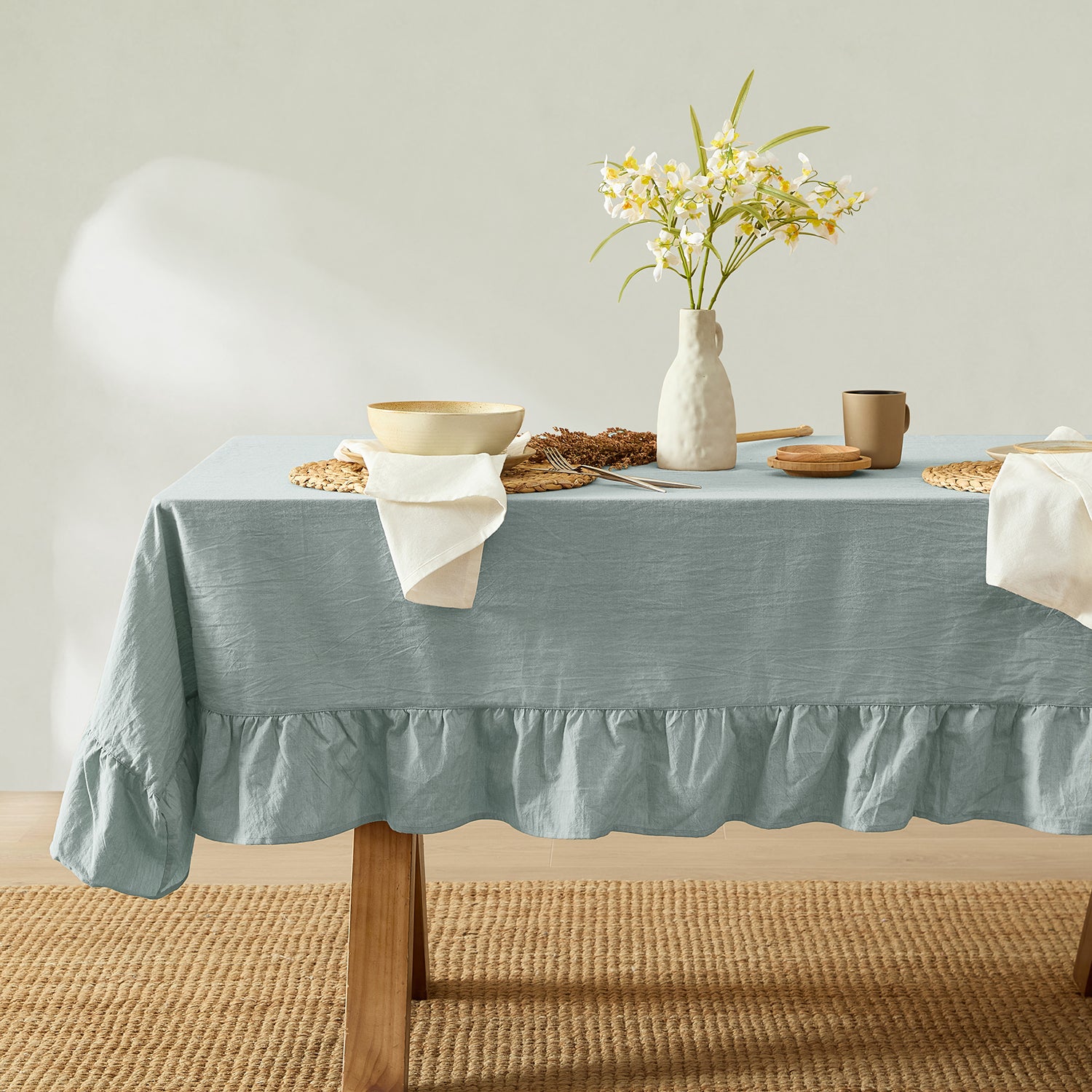 Table setting with a light blue tablecloth, white vase with flowers, and various tableware on a neutral background.