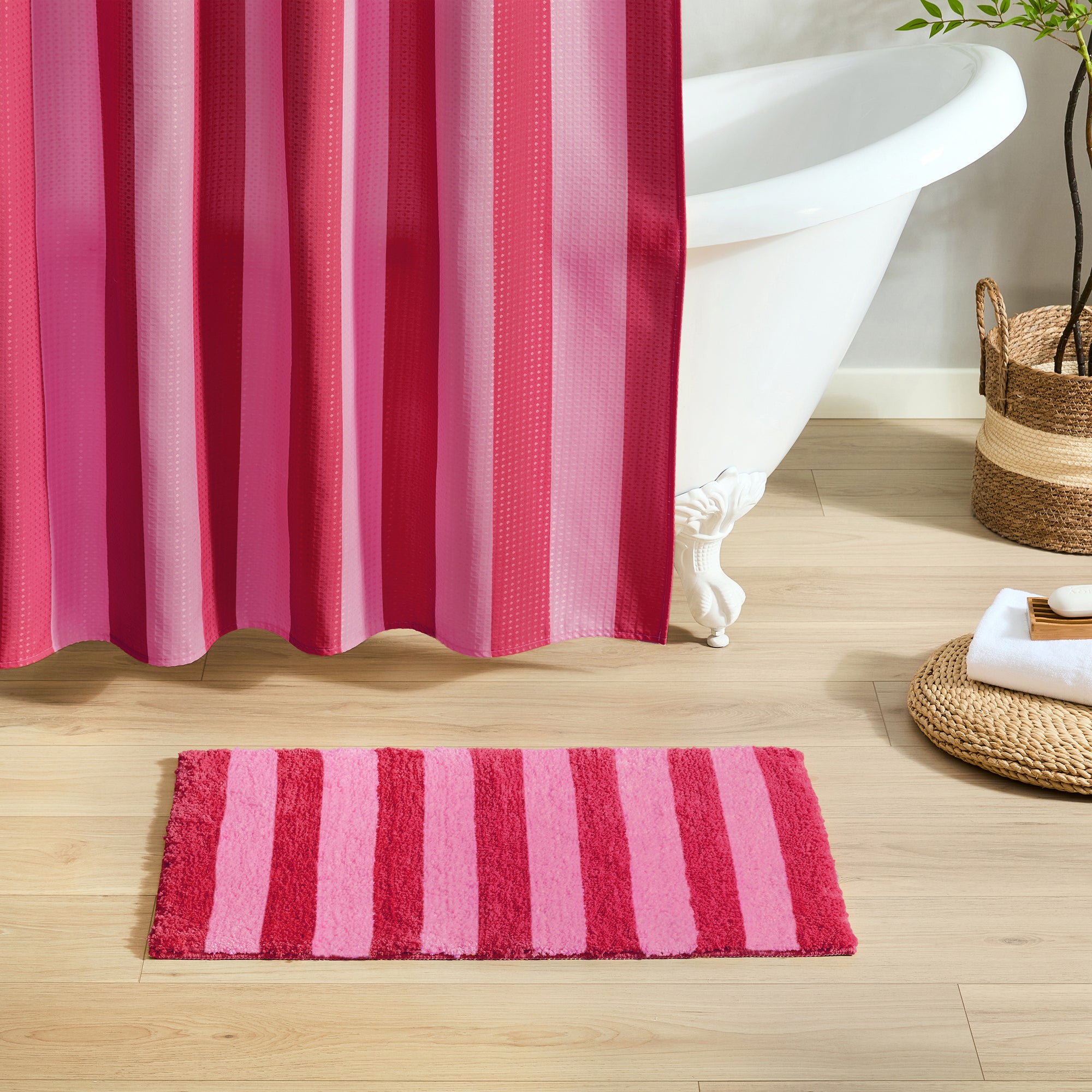 Pink and red striped shower curtain and bath mat on a wooden floor with a bathtub in the background.