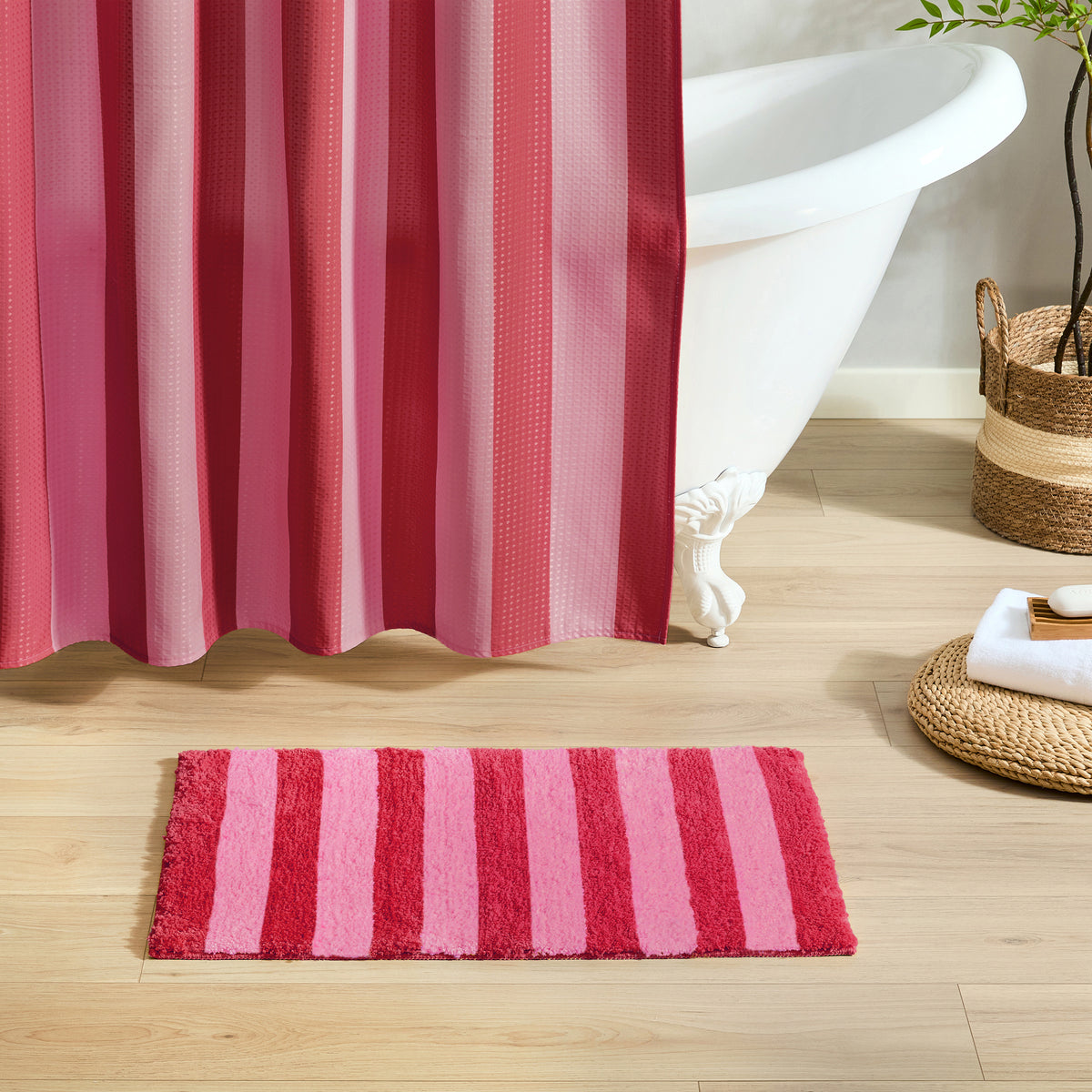 A pink and red striped bath mat on a light wood floor in a bathroom setting, with a white clawfoot tub, a pink striped shower curtain, and woven baskets nearby.