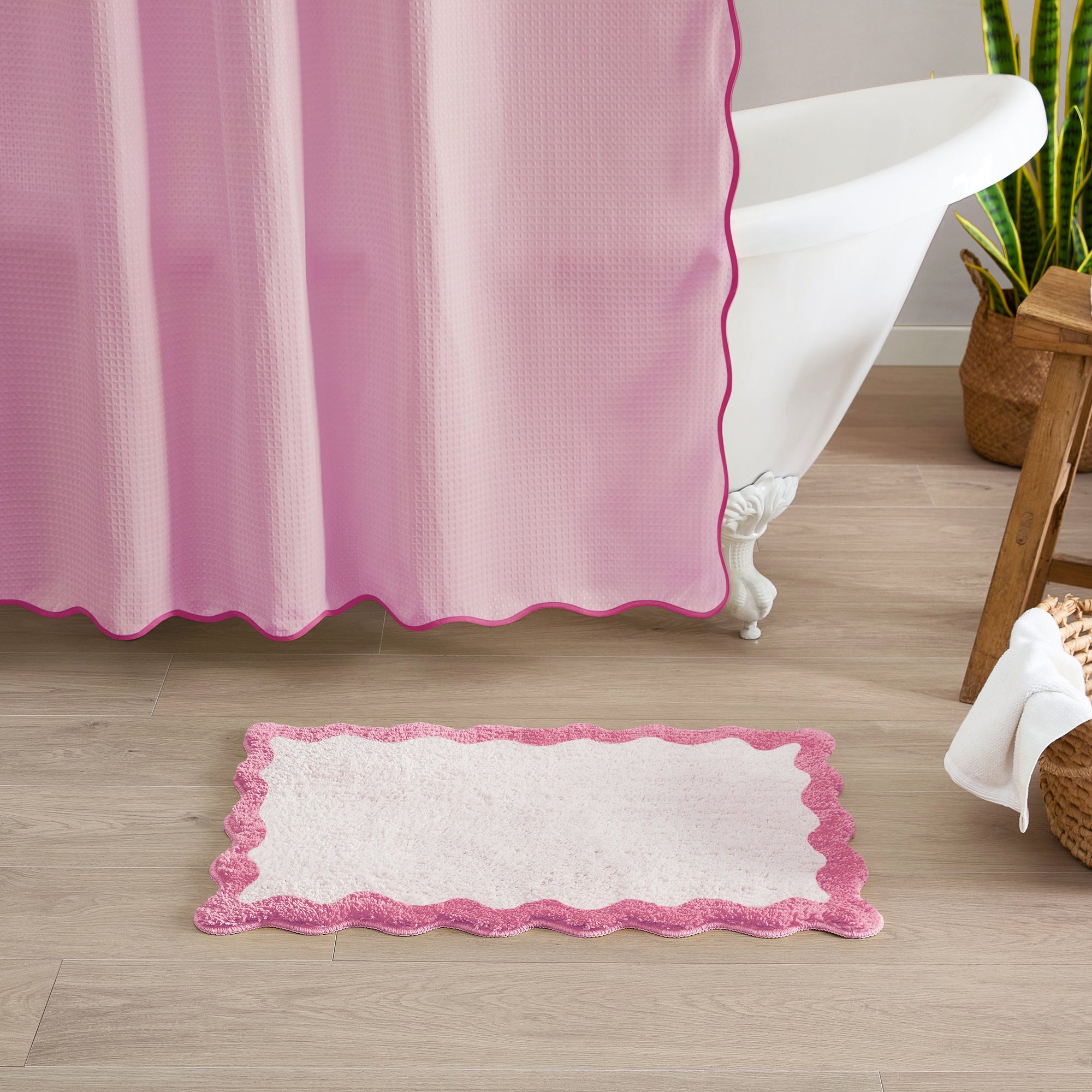 Pink and white bath mat in front of a pink shower curtain on a wooden floor.