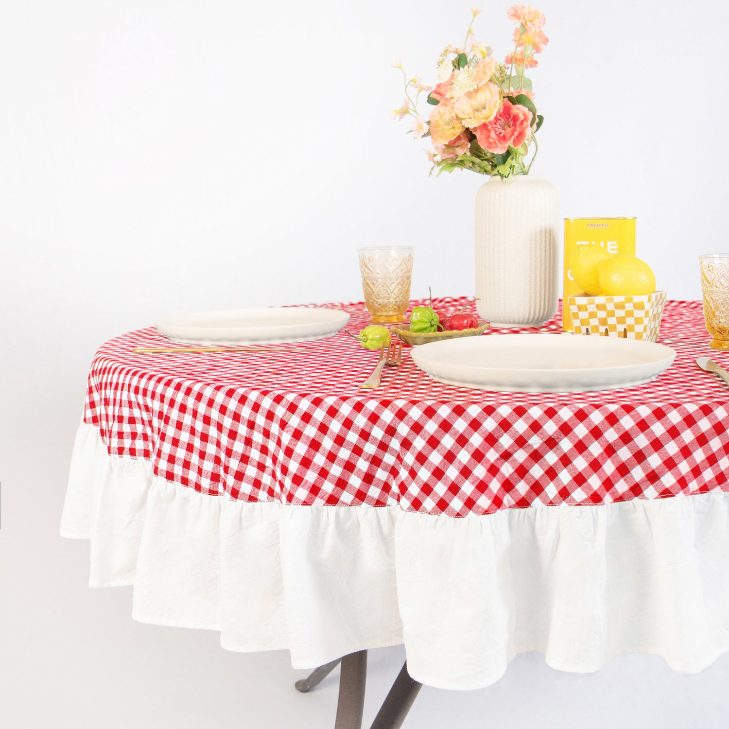 Round table with red and white checkered tablecloth, plates, glasses, and a vase with flowers on a white background.