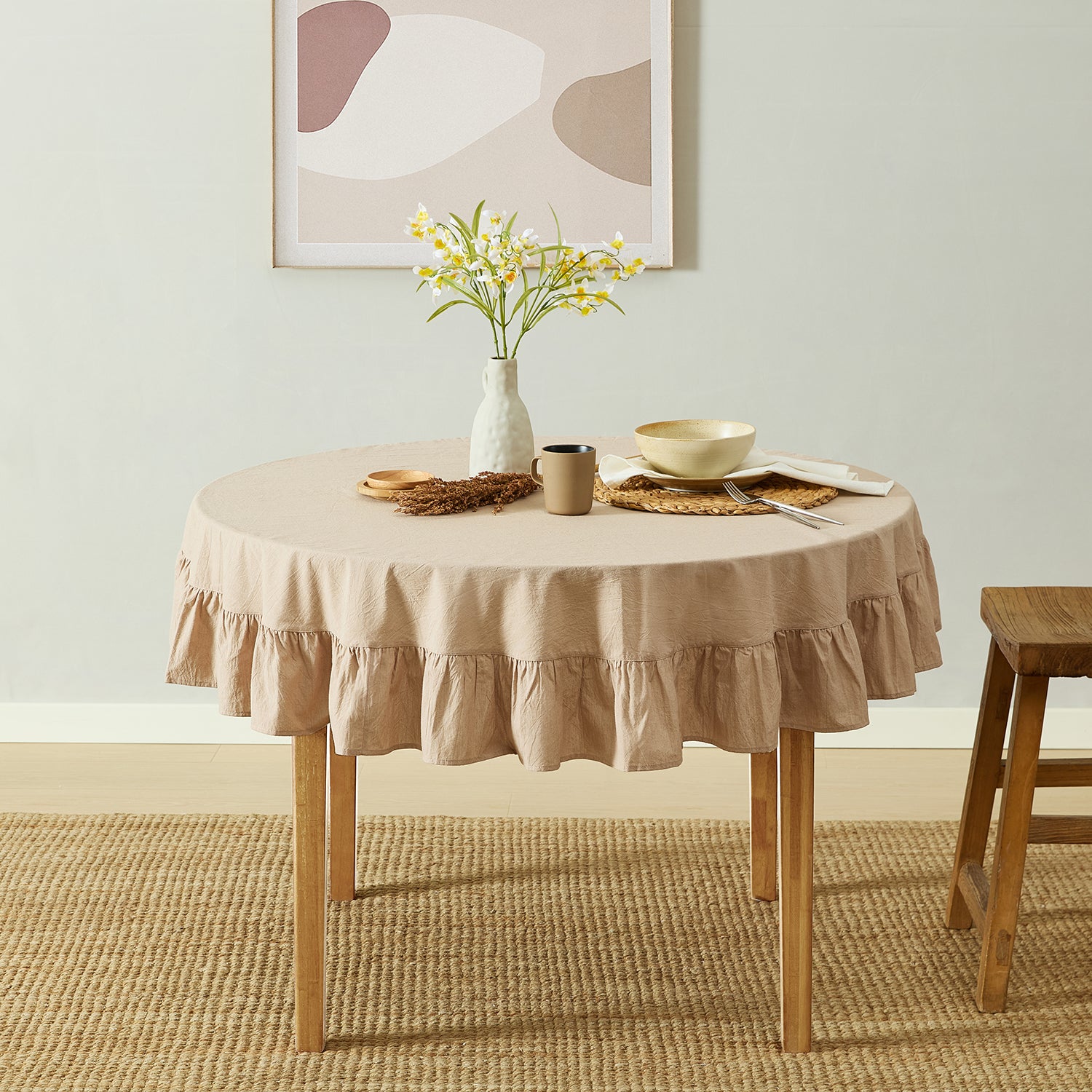 Round table with beige tablecloth, vase with flowers, and wooden stool in a room.