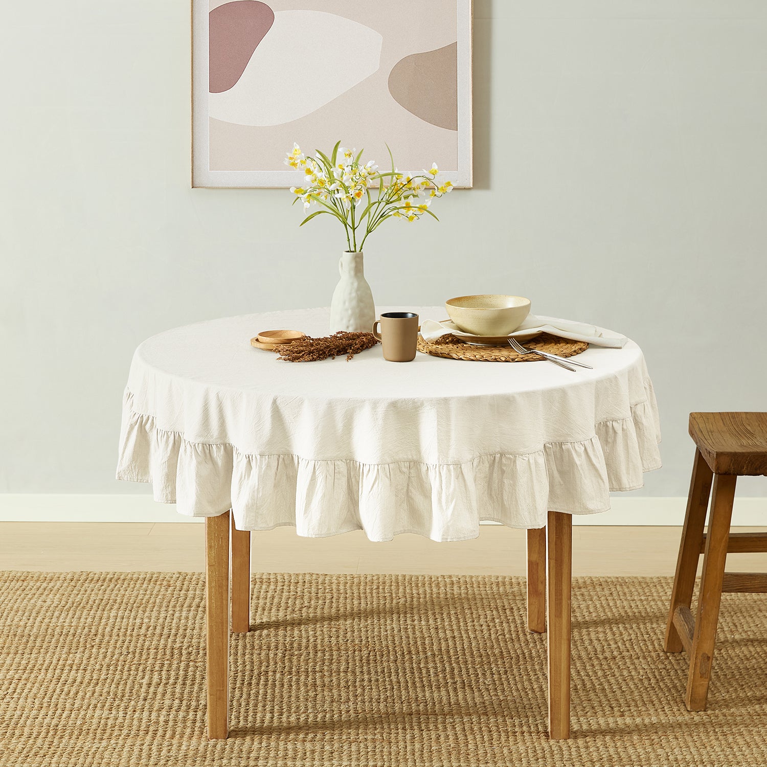Round table with a white tablecloth, vase with flowers, and wooden stool in a room.