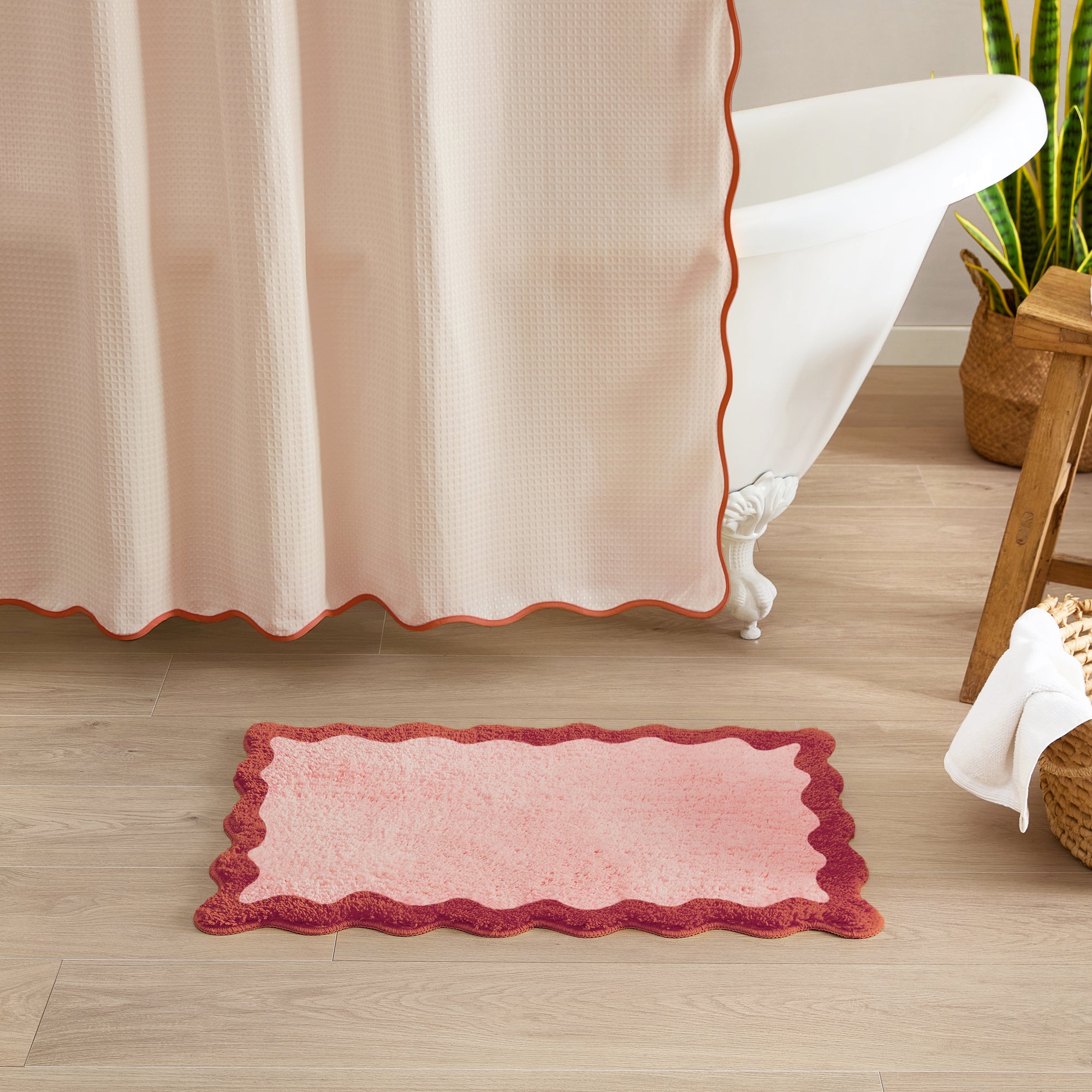 Pink bath mat with scalloped edge on a wooden floor next to a shower curtain.