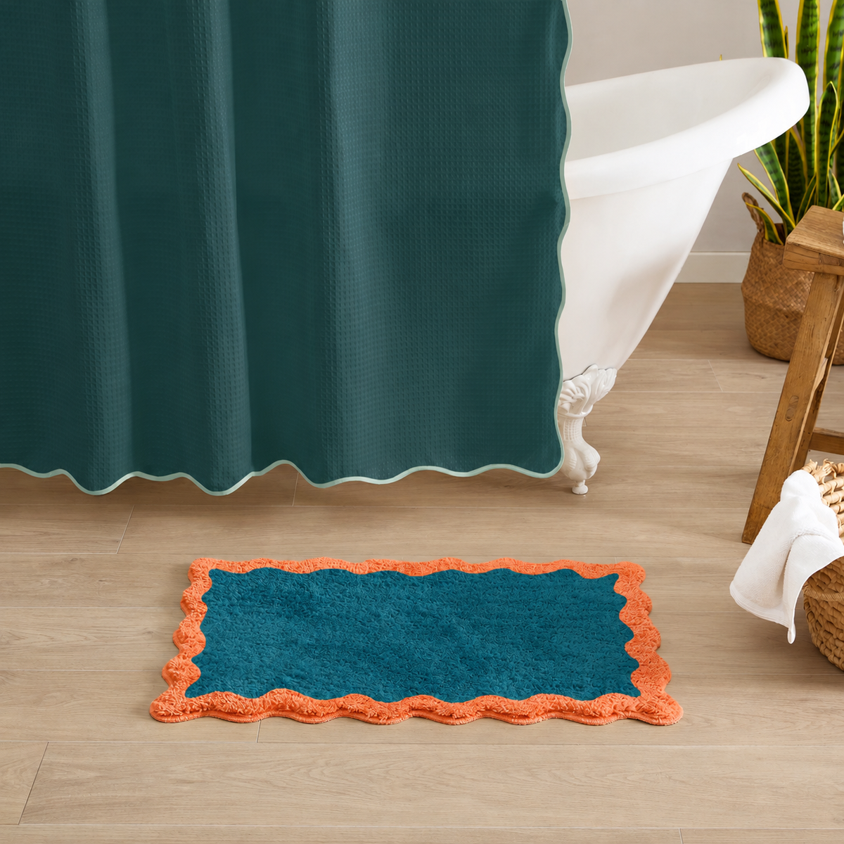 A rectangular bath mat in teal with a bright orange scalloped border, placed on a light wood bathroom floor next to a white clawfoot tub and a dark teal shower curtain.