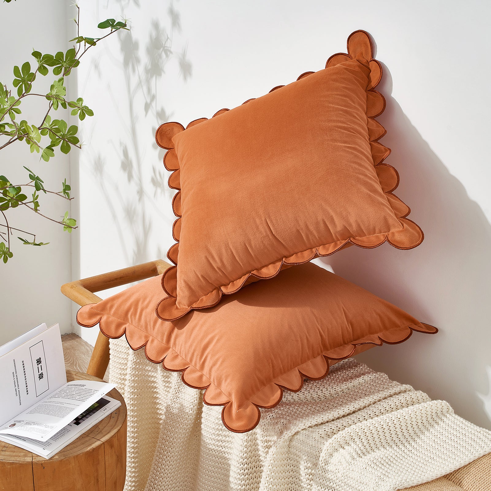 Two orange pillows with scalloped edges on a chair next to a plant and a small table with books.