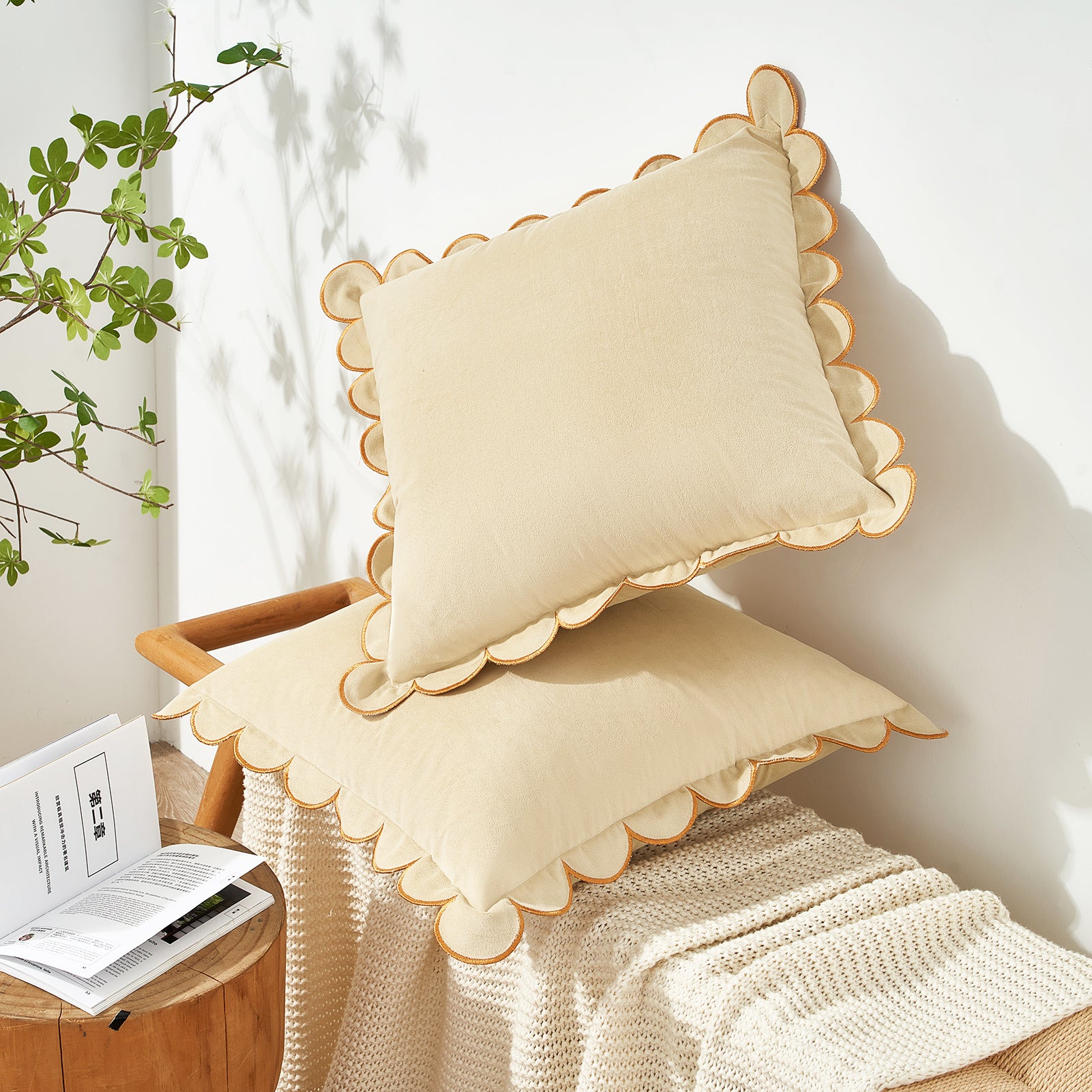Beige pillow with scalloped edge on a wooden chair next to a plant and book.
