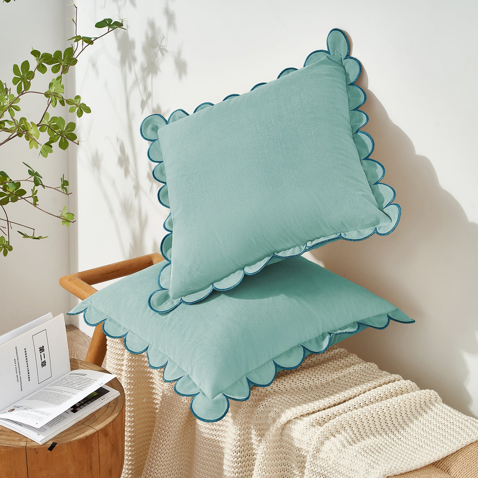 Two light blue pillows with scalloped edges on a beige couch next to a plant and book.