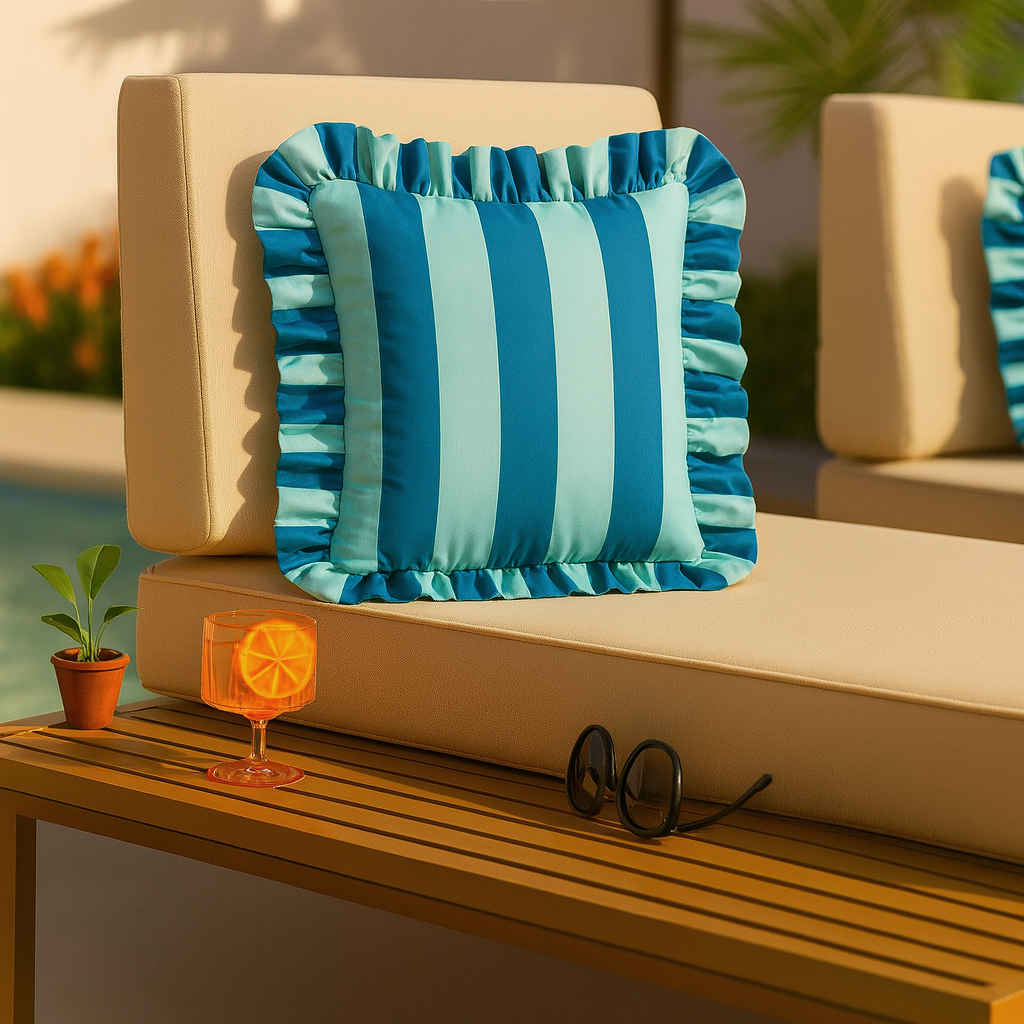 Blue and green striped pillow on a beige outdoor cushion with a drink and sunglasses on a wooden table.