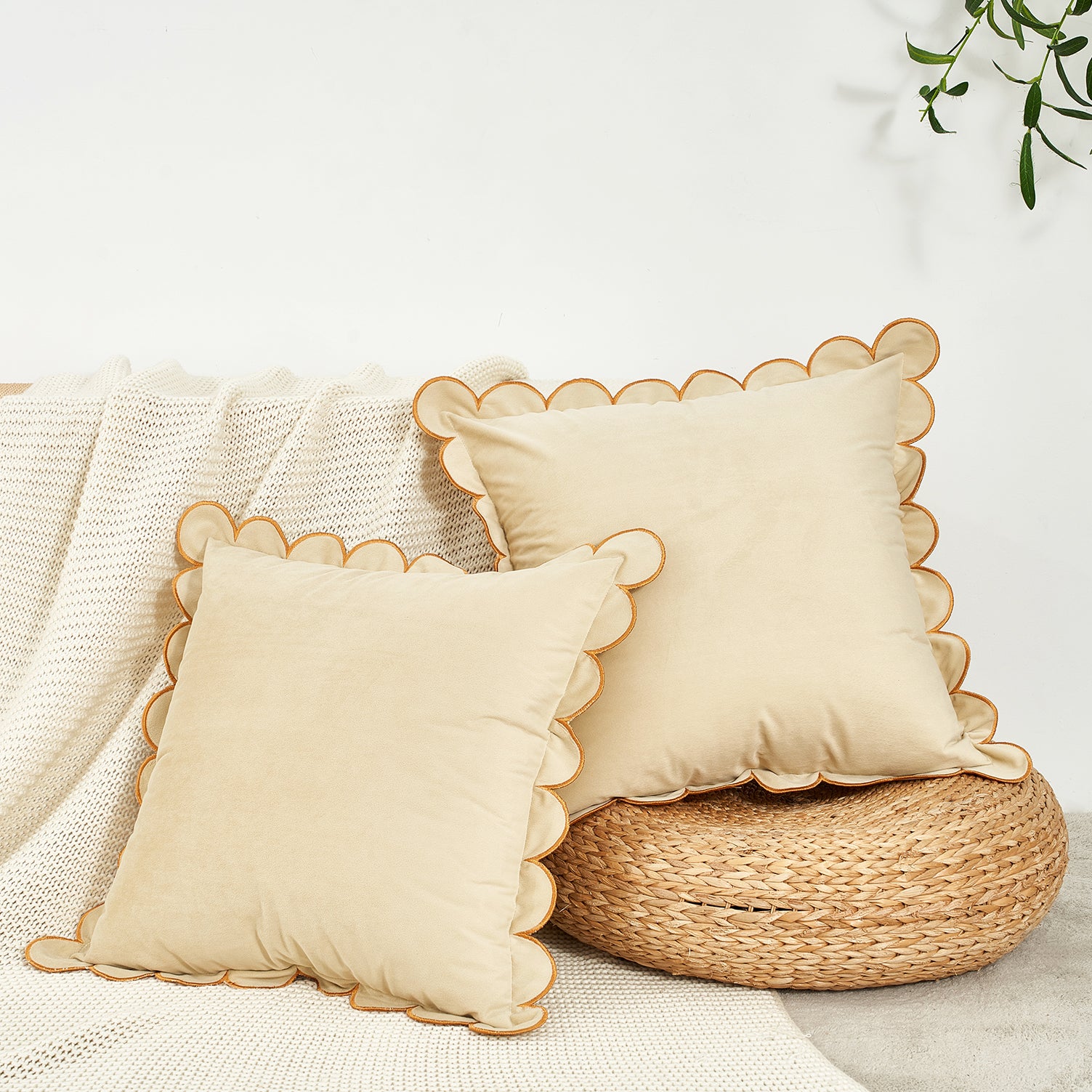 Two beige pillows with scalloped edges on a woven stool against a white background.