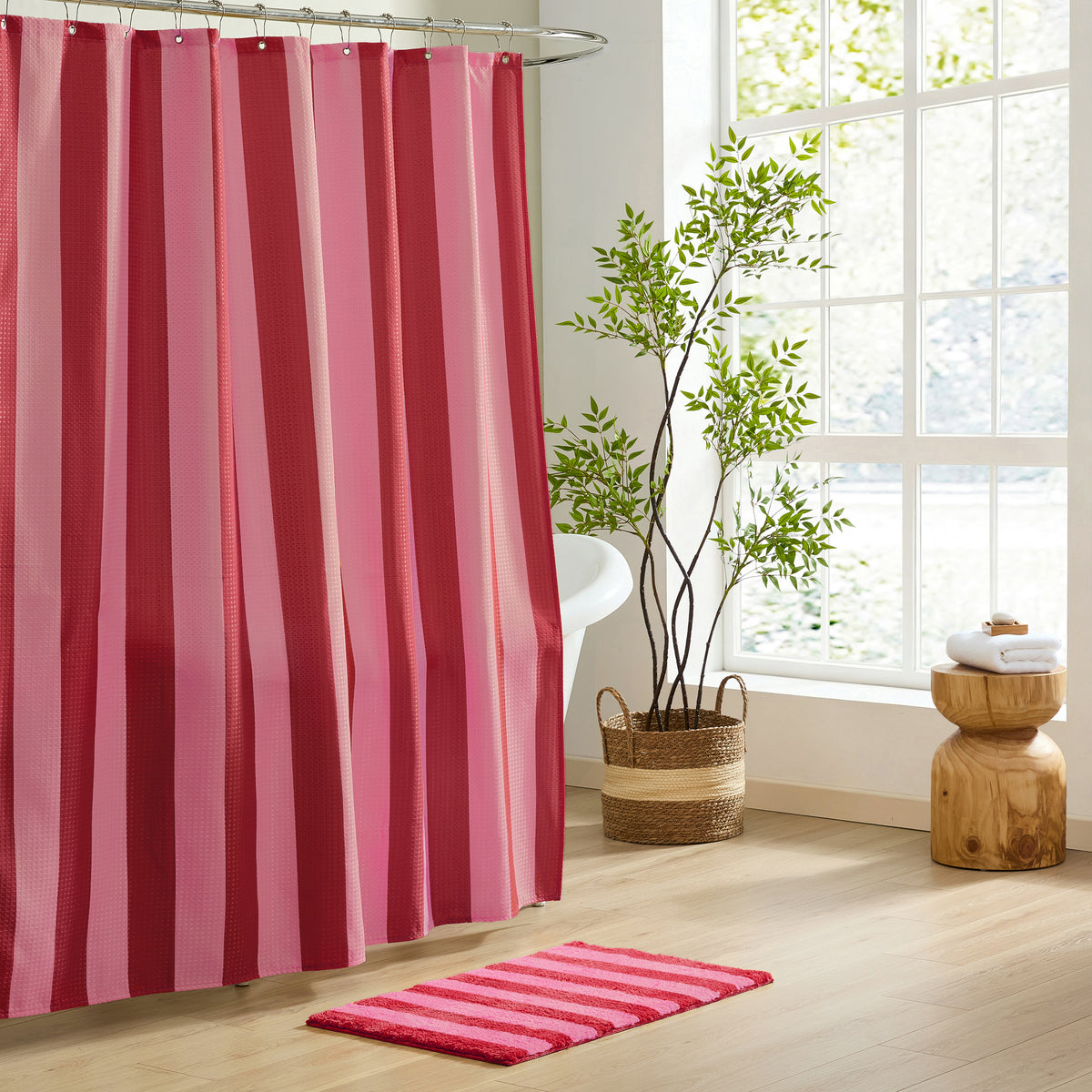 A pink and red vertically striped shower curtain hanging in a bright bathroom next to a window, with a matching bath mat on the floor and a potted plant nearby.