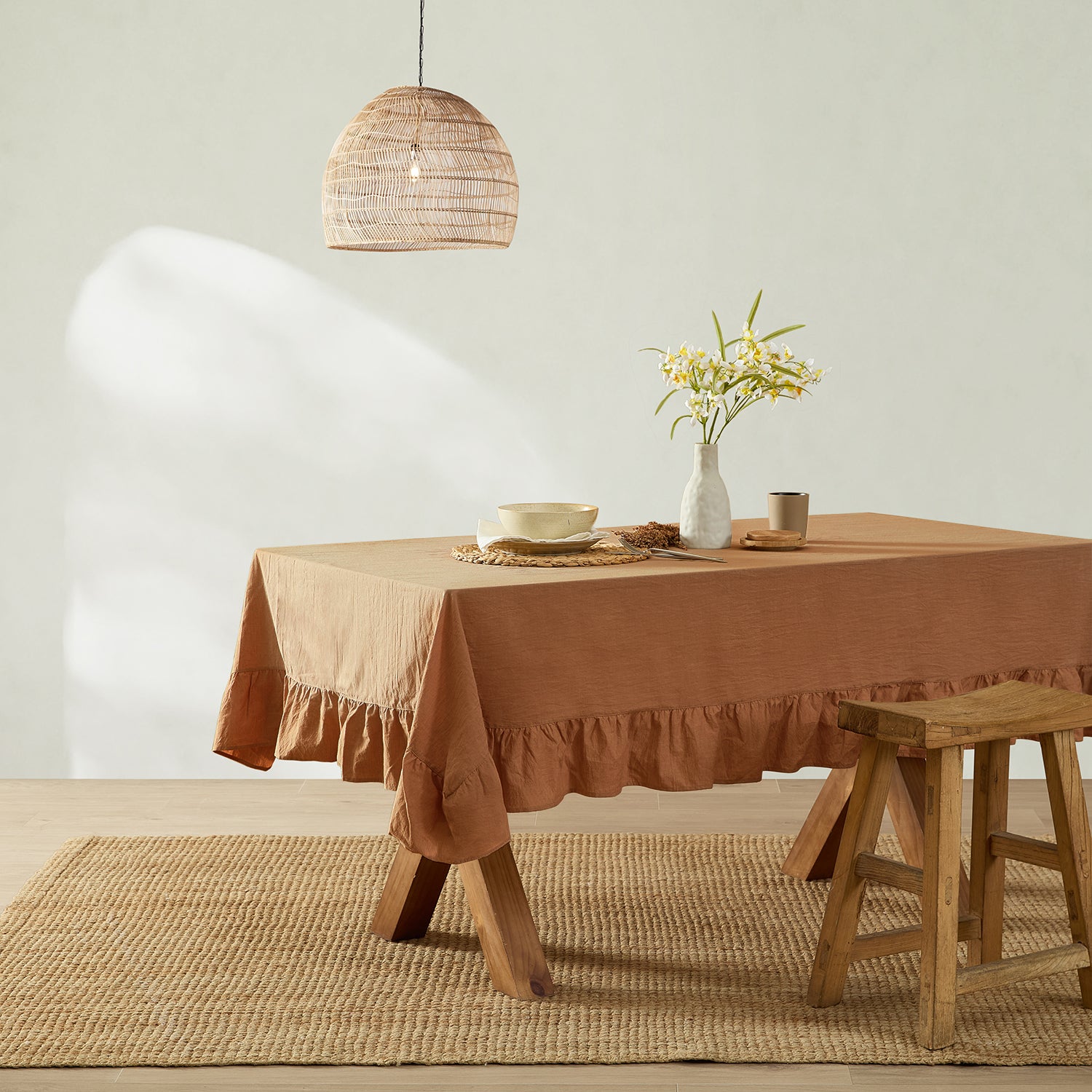 Dining table with a brown tablecloth, vase with flowers, and wooden stool in a room with a light-colored wall and pendant lamp.