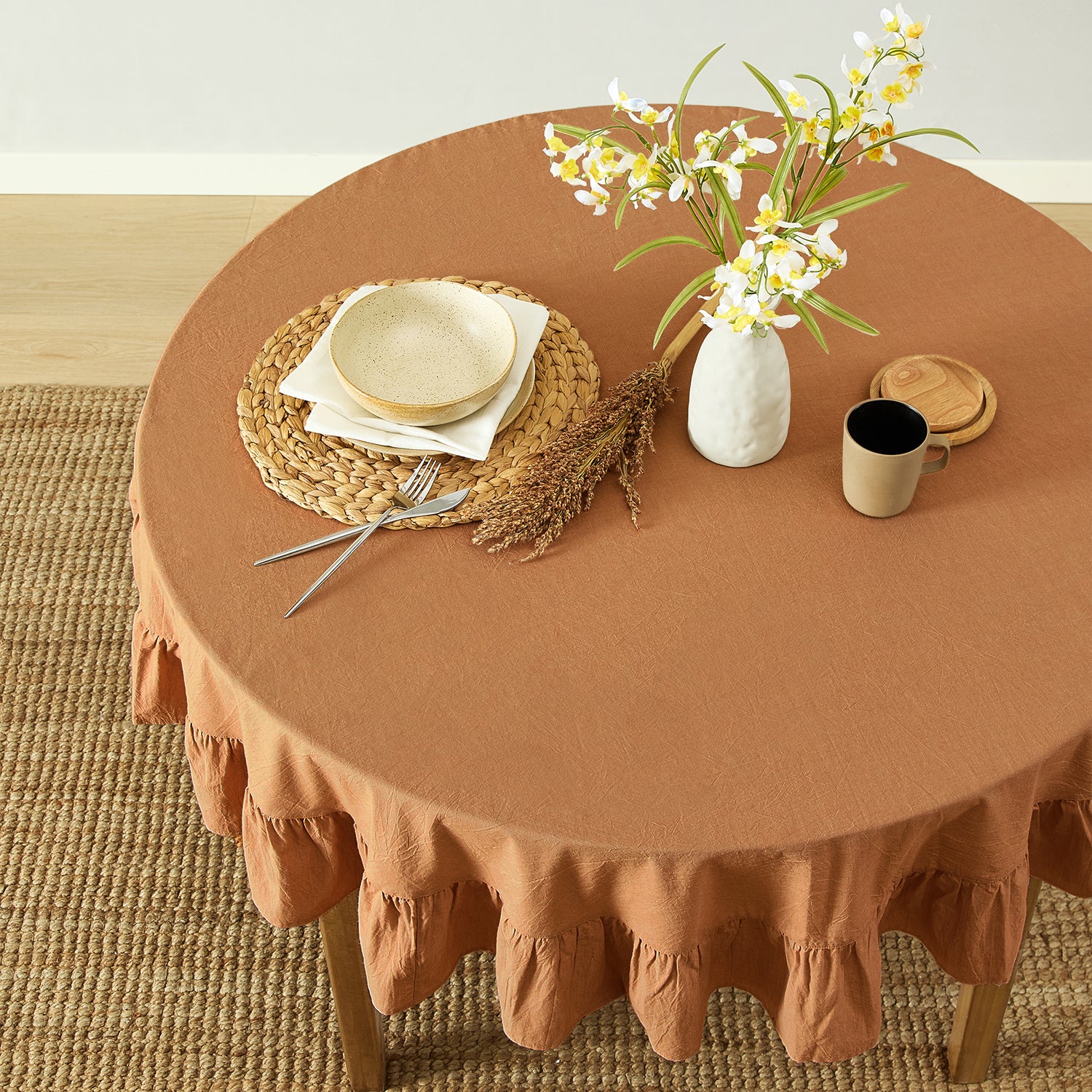 Round table with a brown tablecloth, plates, cutlery, and a vase with flowers on a wooden floor.