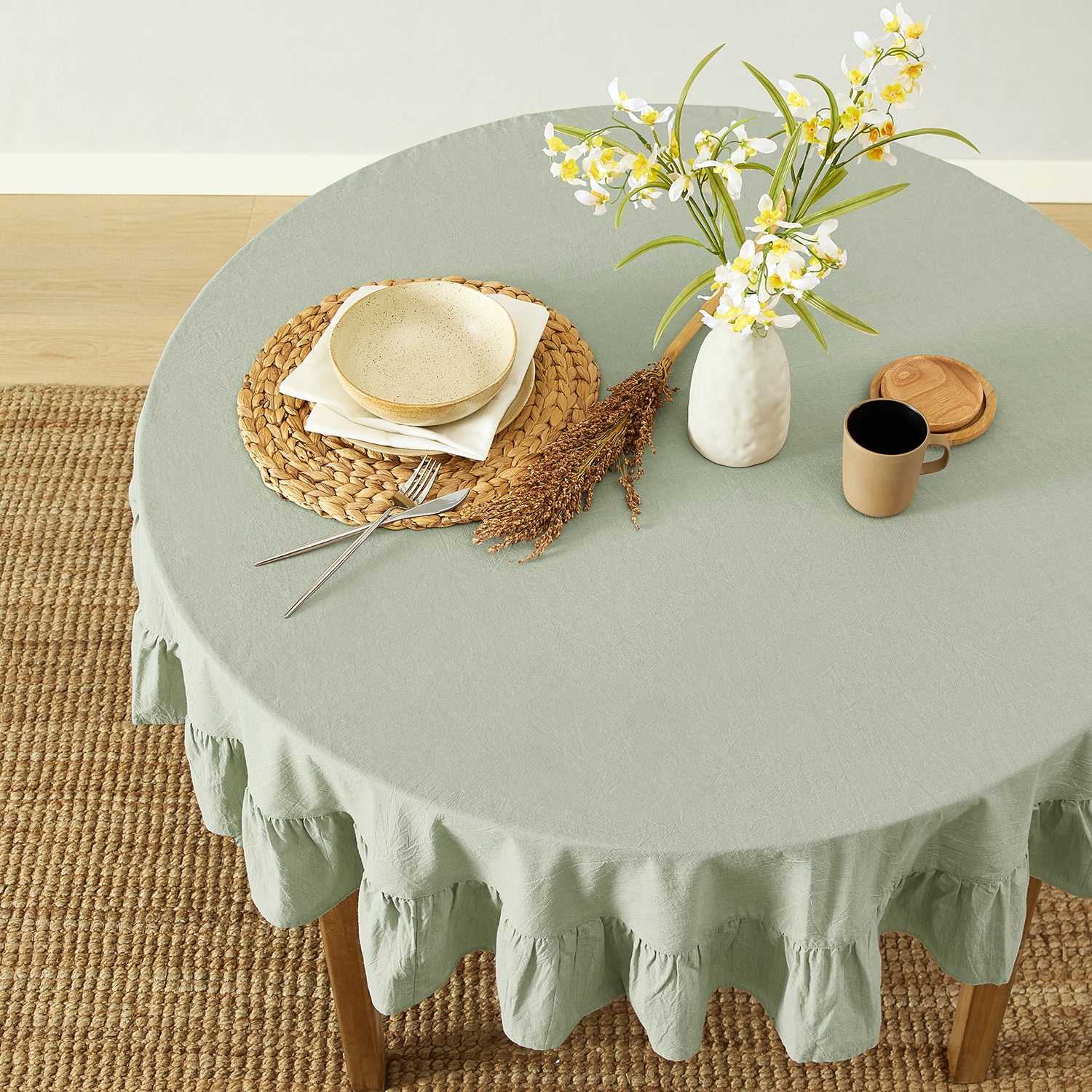 Round table with a green tablecloth, plates, cutlery, and a vase with flowers on a wooden floor.