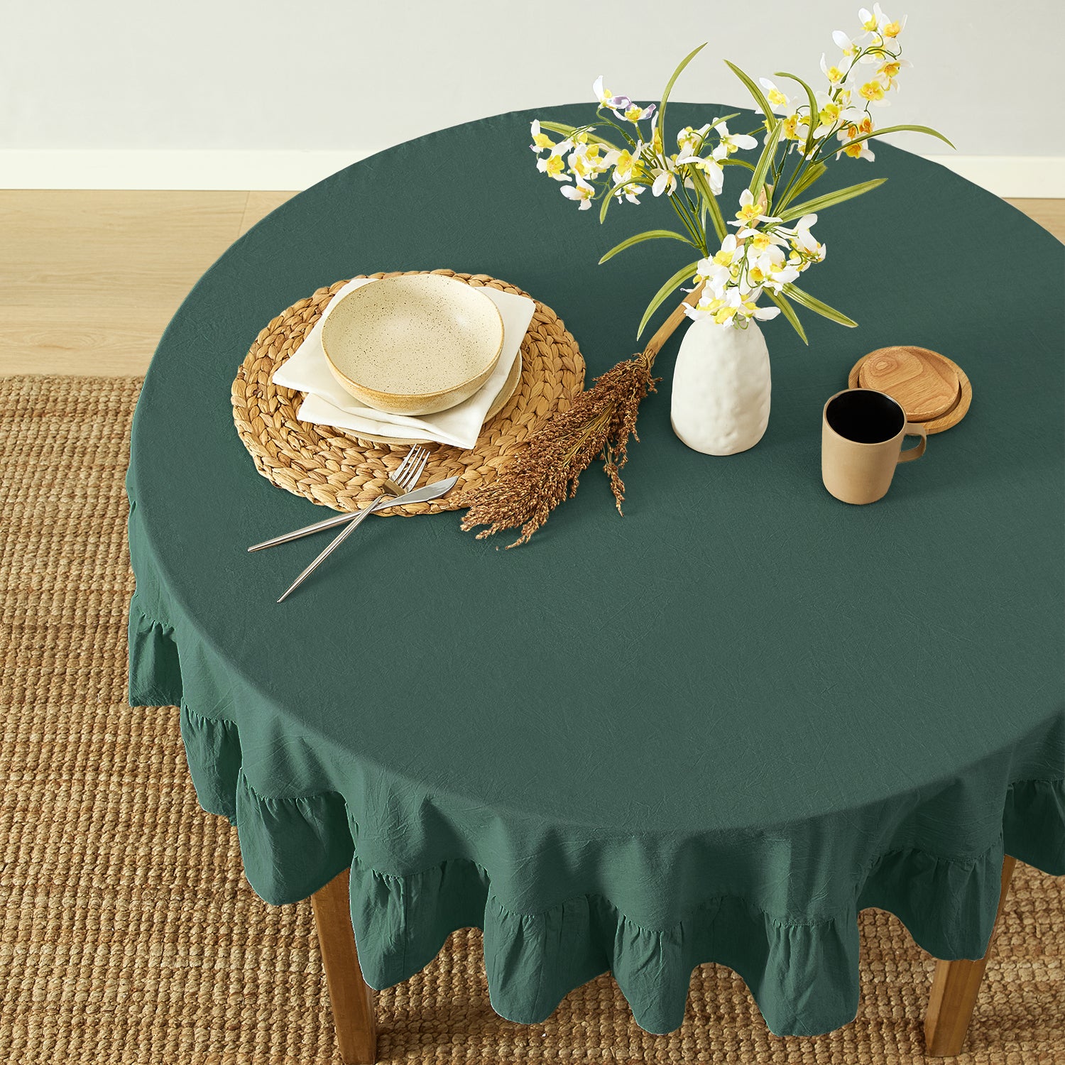 Round table with a green tablecloth, plates, cutlery, and a vase with flowers on a wooden floor.