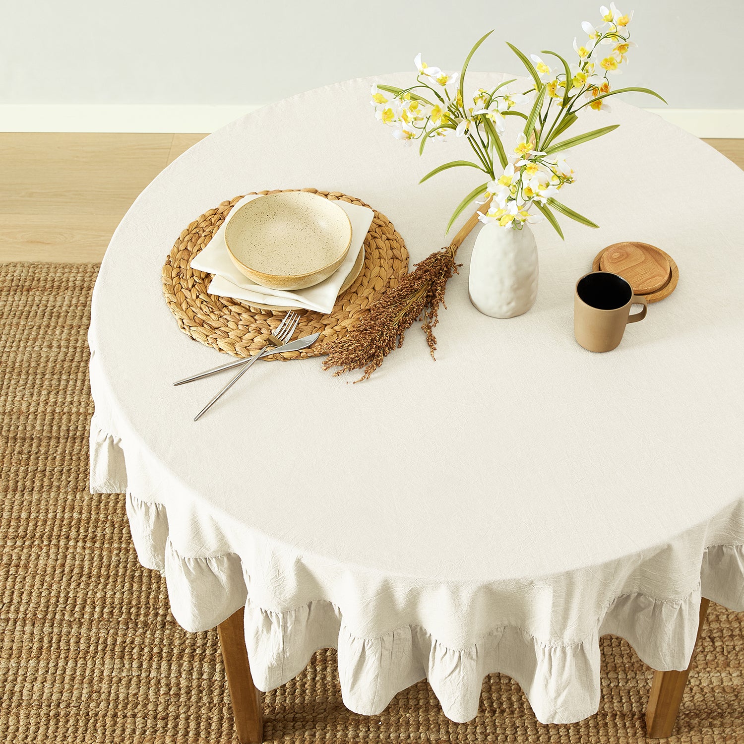 Round table with a white tablecloth, plates, cutlery, and a vase with flowers on a wooden floor.