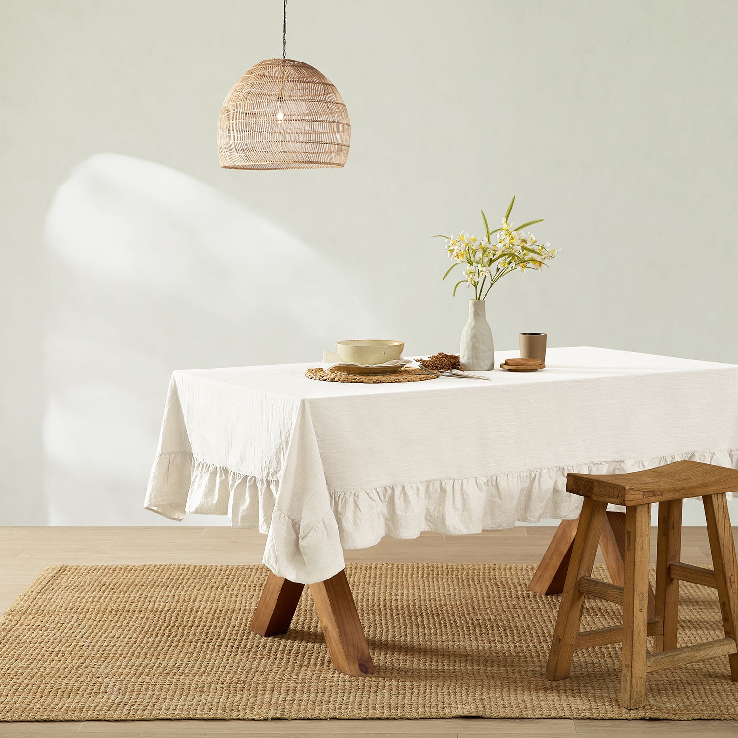 Dining room with a table set with a white tablecloth, bowls, and a vase with flowers, wooden stool, and pendant light.