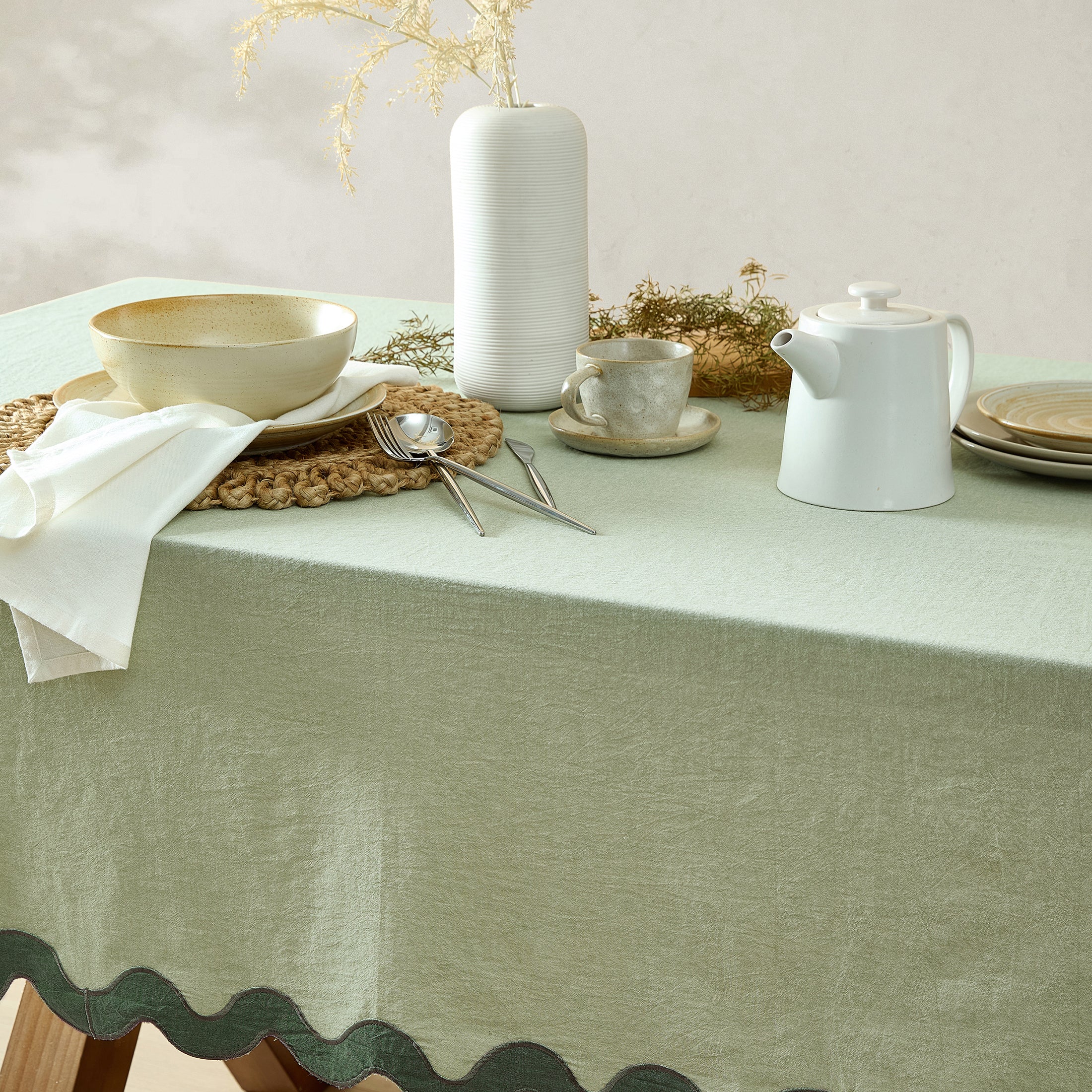Dining table setting with a green scalloped tablecloth, white teapot, cup, and utensils.