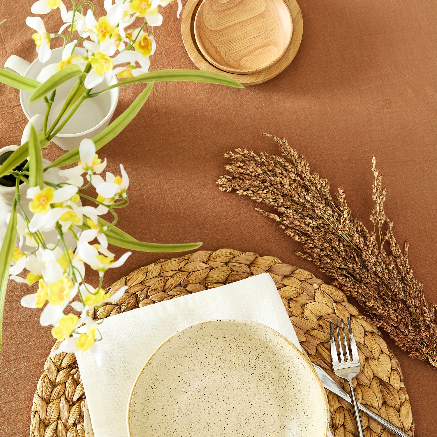 Table setting with ceramic plates, woven placemat, flowers, and cutlery on a brown tablecloth.