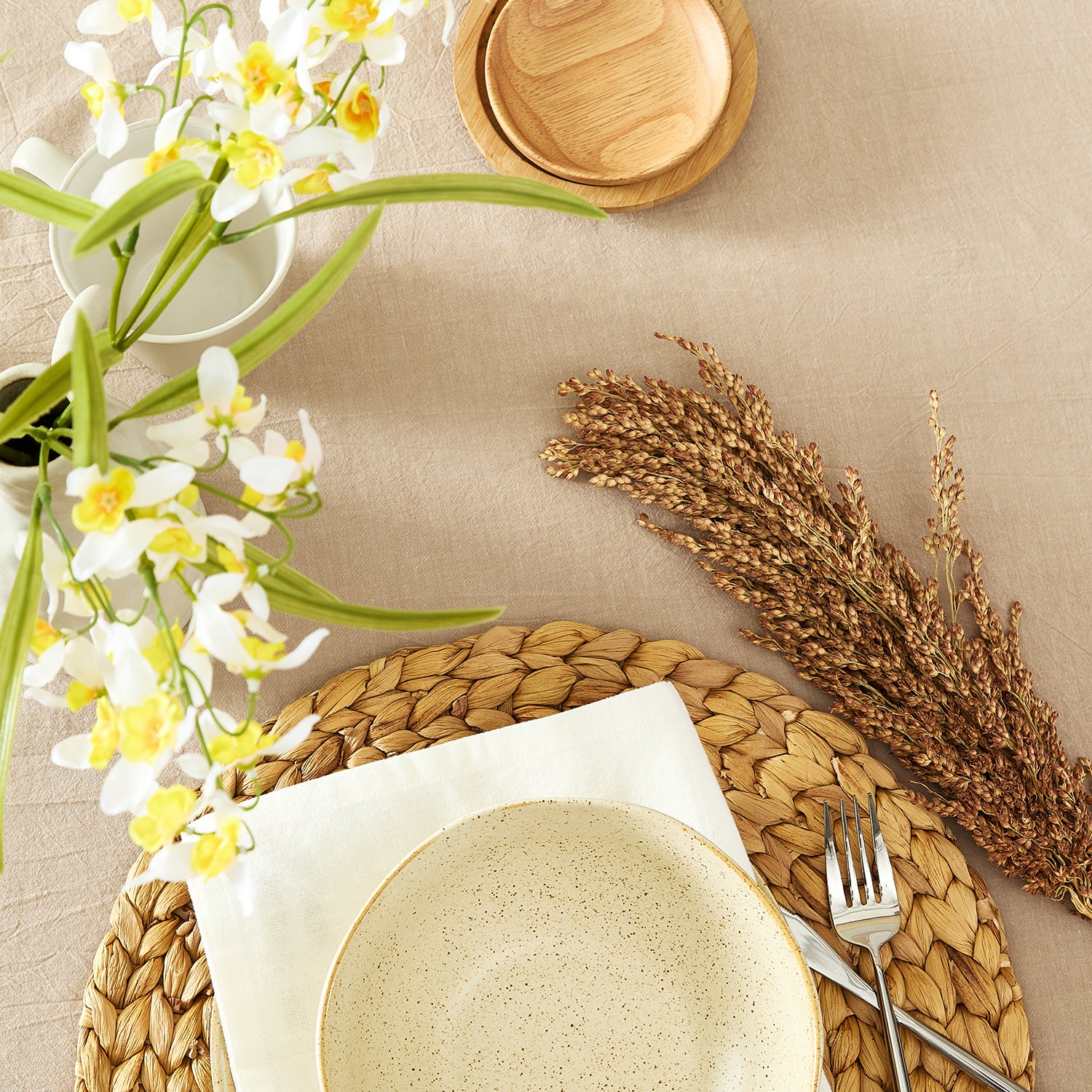 Table setting with woven placemat, ceramic plate, flowers, and wheat on a beige surface.