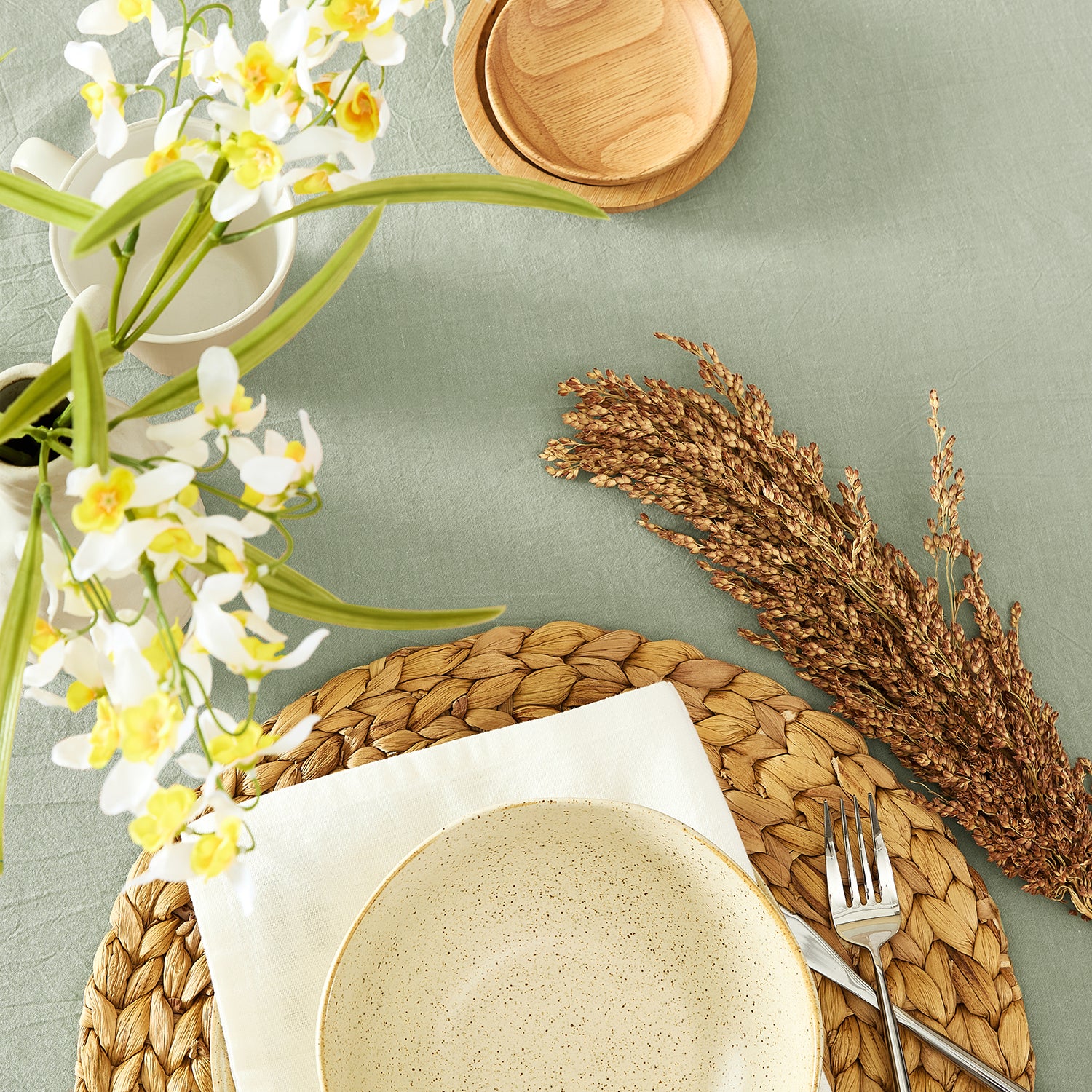 Table setting with ceramic plates, woven placemat, flowers, and wheat on a light green background