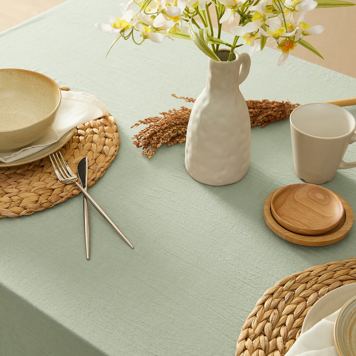 Table setting with ceramic plates, cutlery, a vase, and flowers on a light green tablecloth.