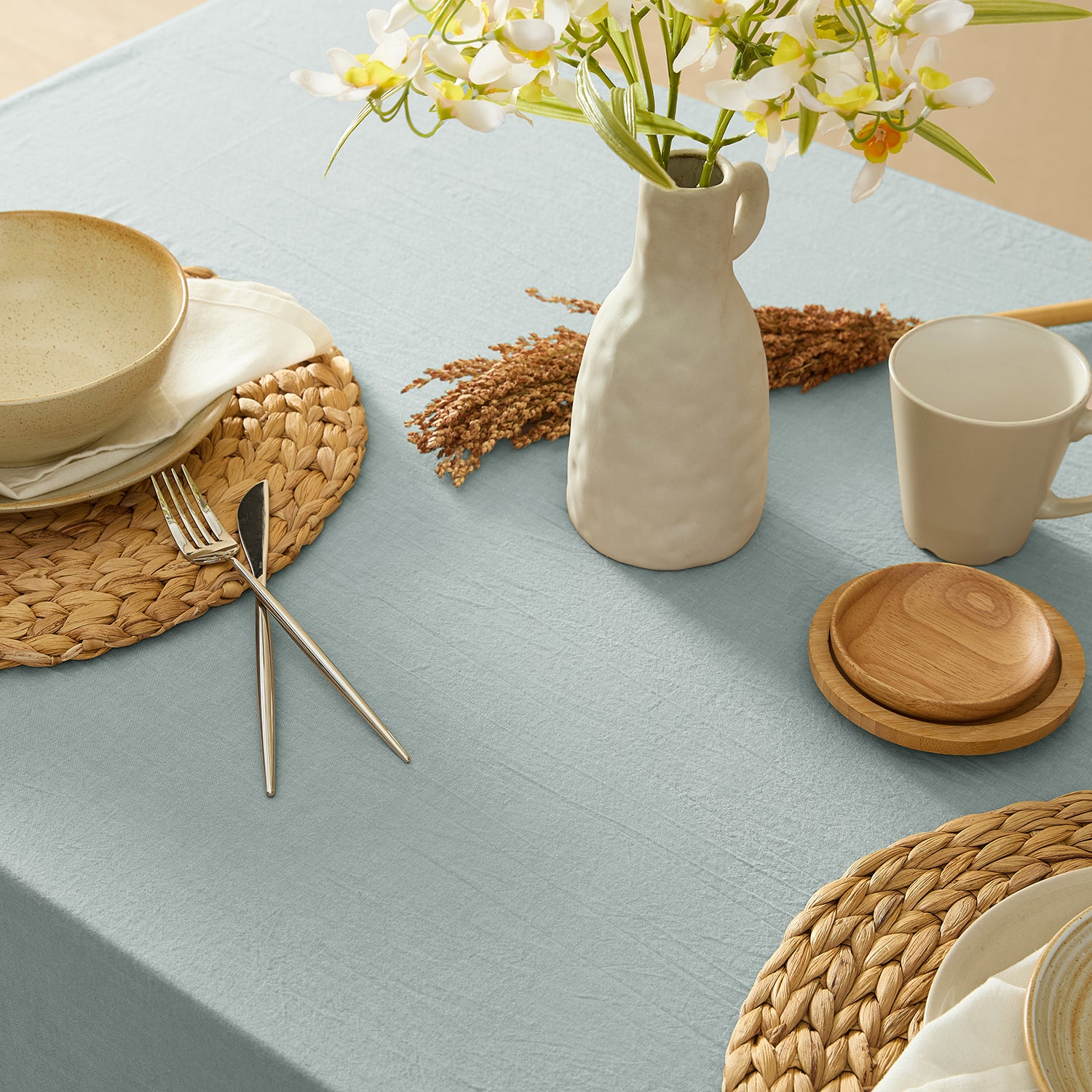 Table setting with ceramic plates, cutlery, a vase, and flowers on a light blue tablecloth.