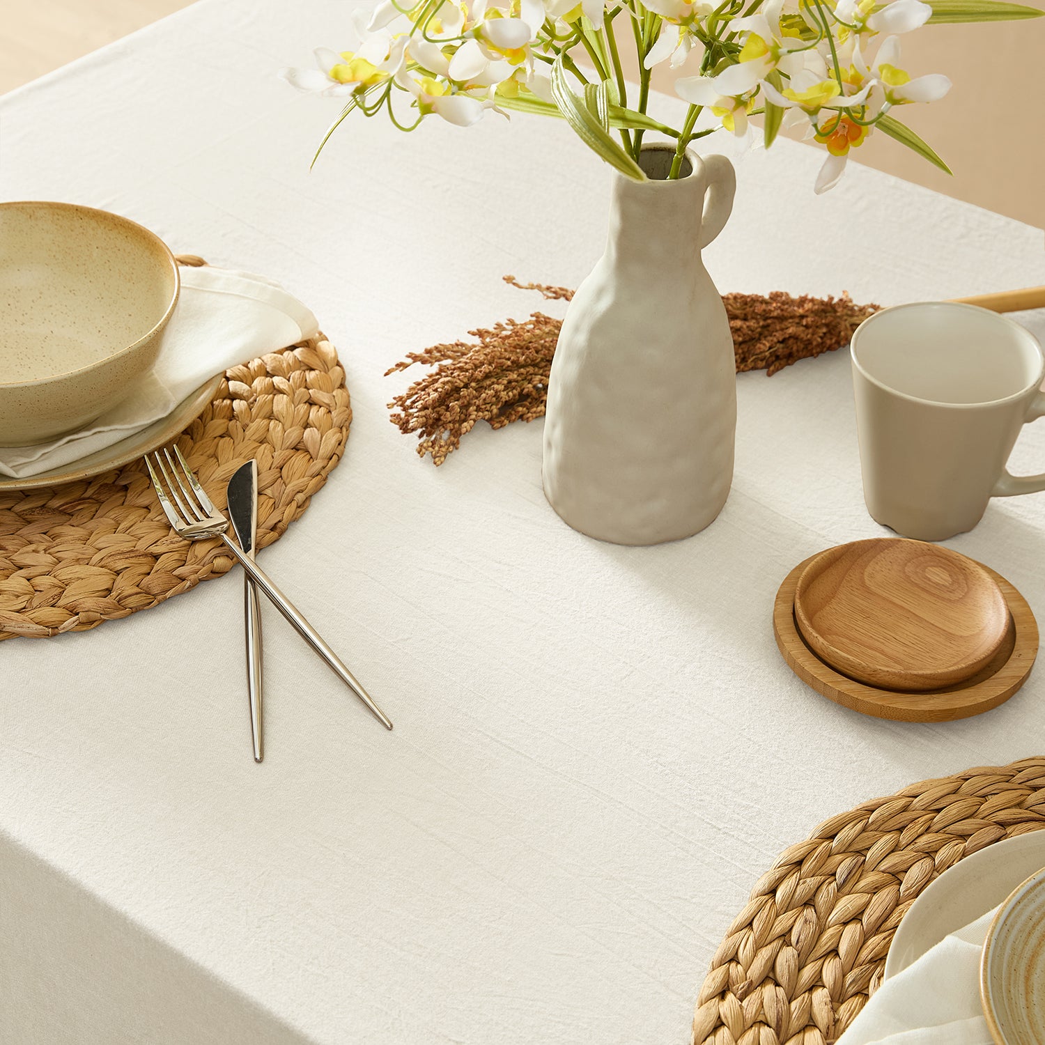 Table setting with ceramic plates, wooden coasters, and a vase with flowers on a white tablecloth.