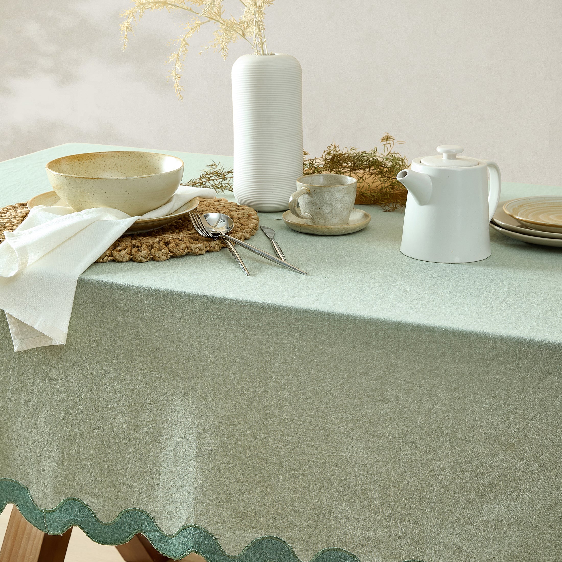 Table setting with a green tablecloth, white teapot, cup, and utensils on a light background.