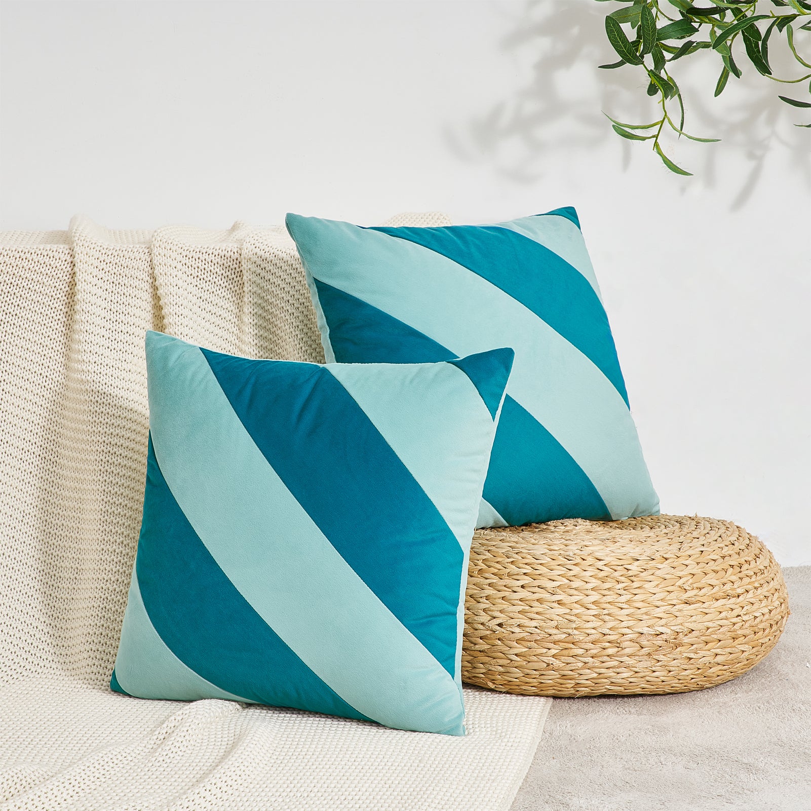 Two blue and green striped pillows on a beige couch with a woven footstool and plant in the background.