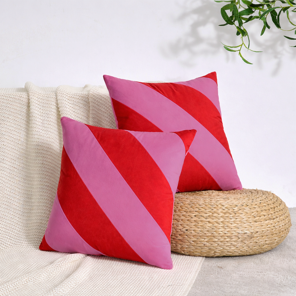Two square throw pillows with diagonal red and pink stripes, styled on a cream knit blanket next to a woven round pouf.