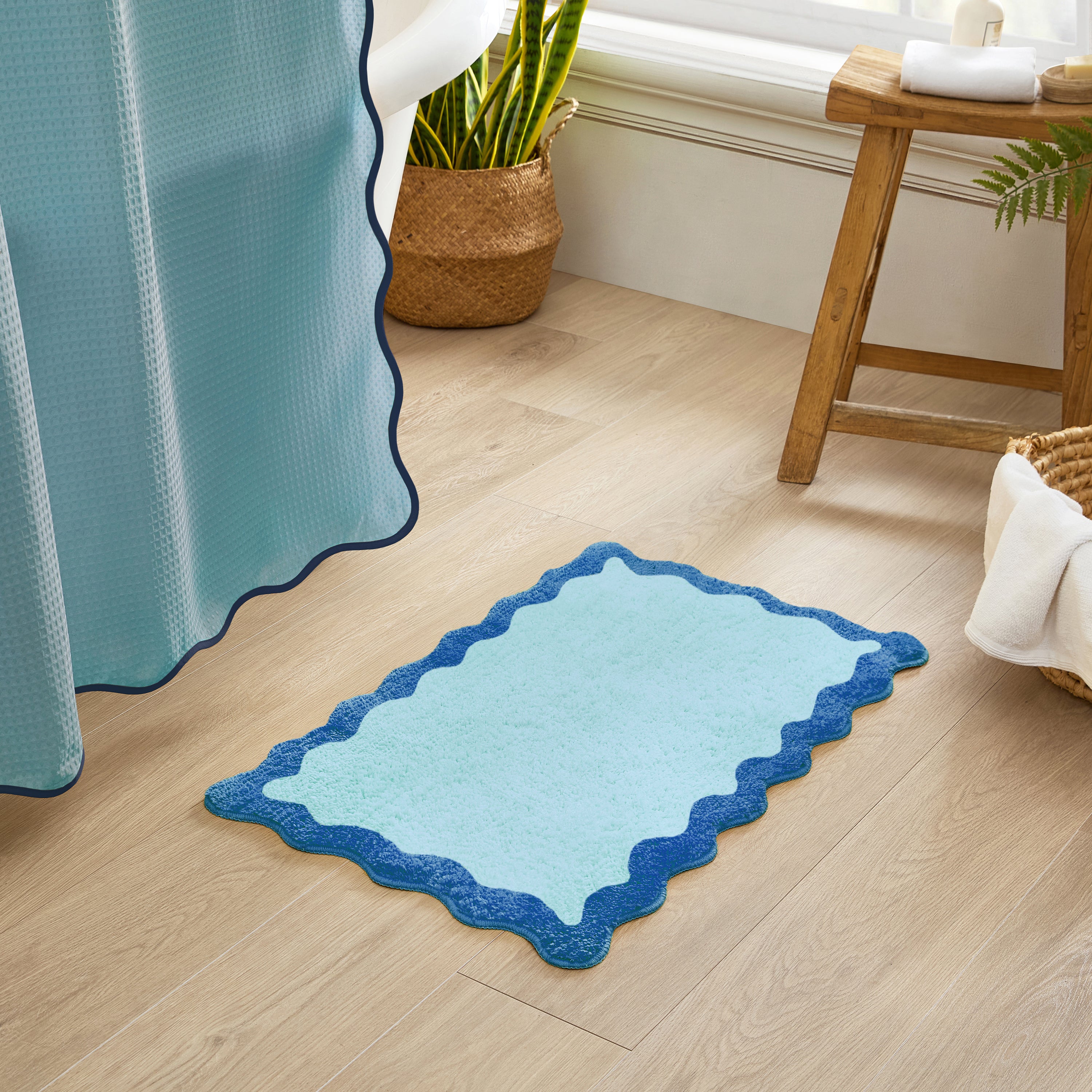 Blue bath mat with scalloped edge on a wooden floor next to a shower curtain.