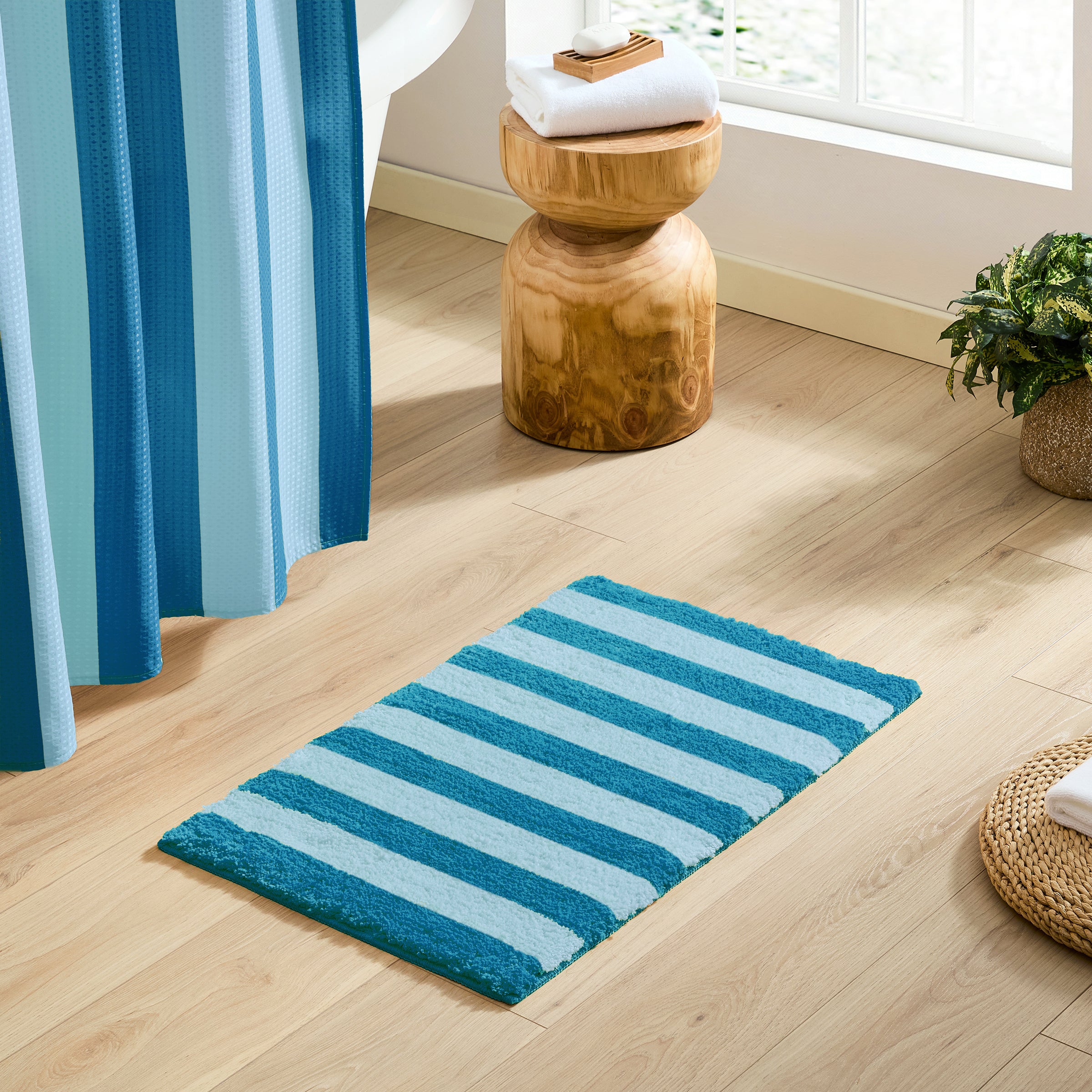 Blue and green striped bath mat on a wooden floor with a wooden stool and plant in the background.