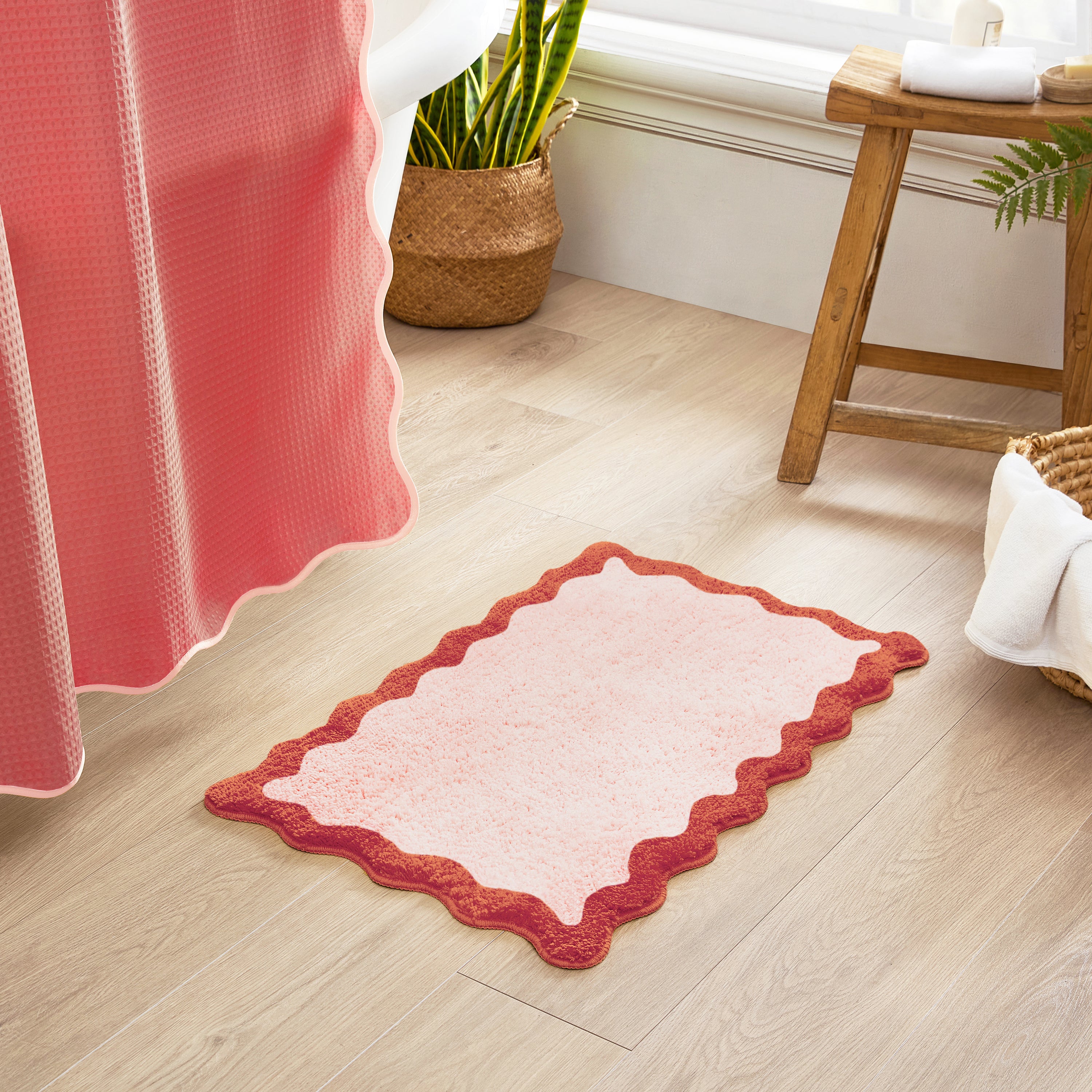 Pink bath mat with scalloped edge on a wooden floor next to a pink shower curtain.
