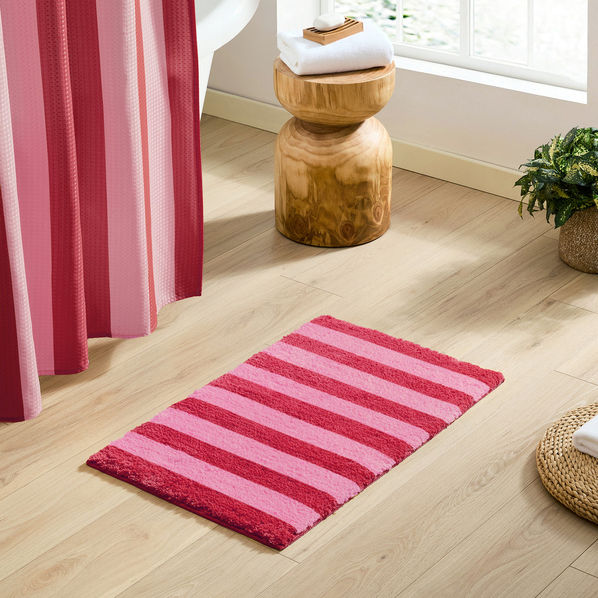 A rectangular bath mat with alternating pink and red stripes lies on a light wood floor in a bright bathroom setting, next to a pink striped shower curtain, a wooden stool, and a potted plant.