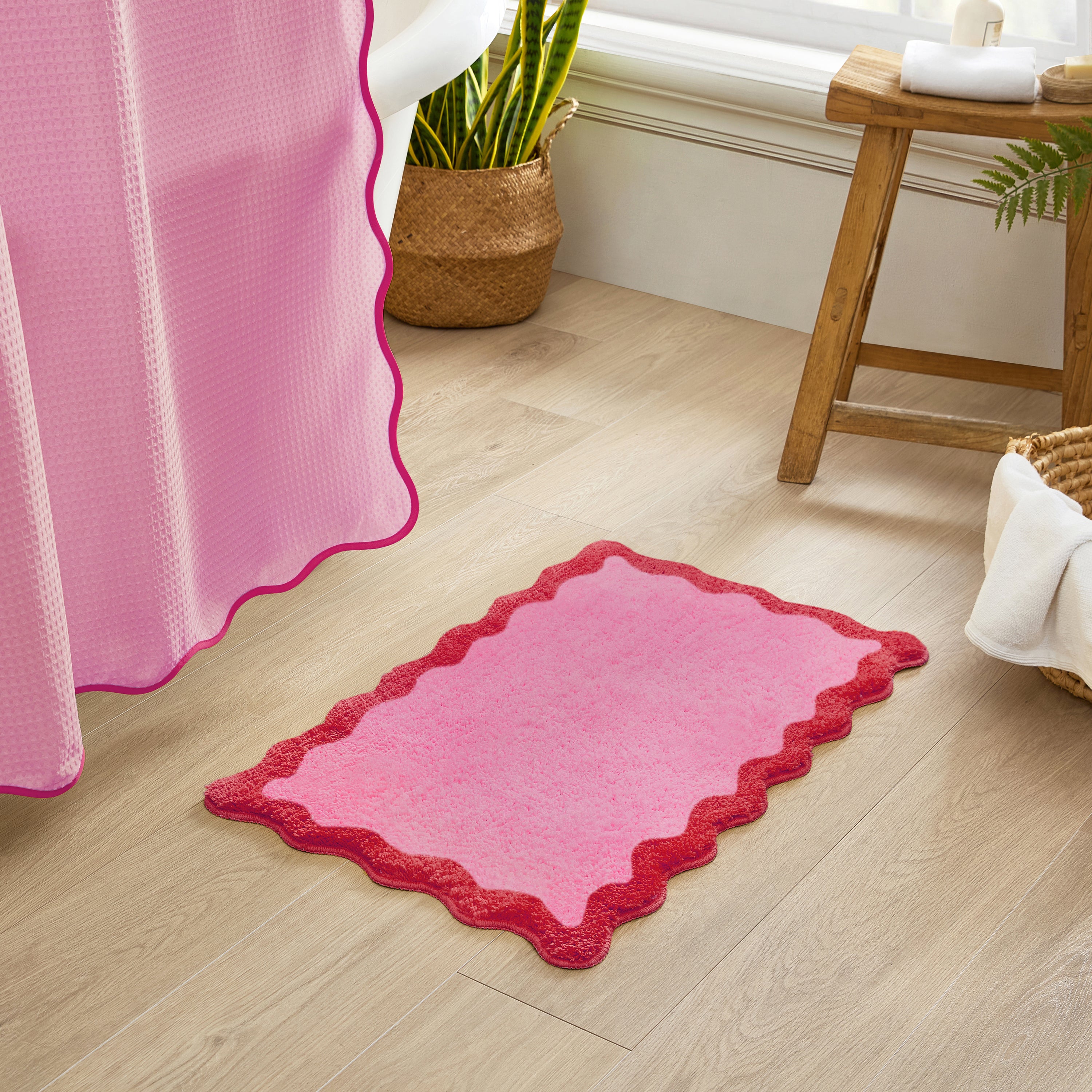 Pink bath mat with scalloped edge on a wooden floor next to a pink shower curtain.