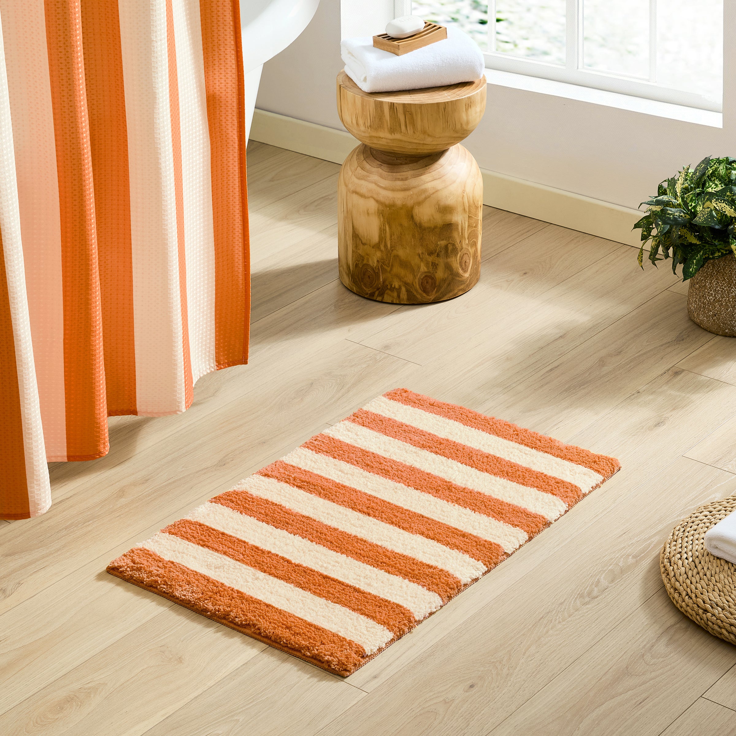 Striped orange and beige bath mat on a wooden floor with a wooden stool and plant in the background.